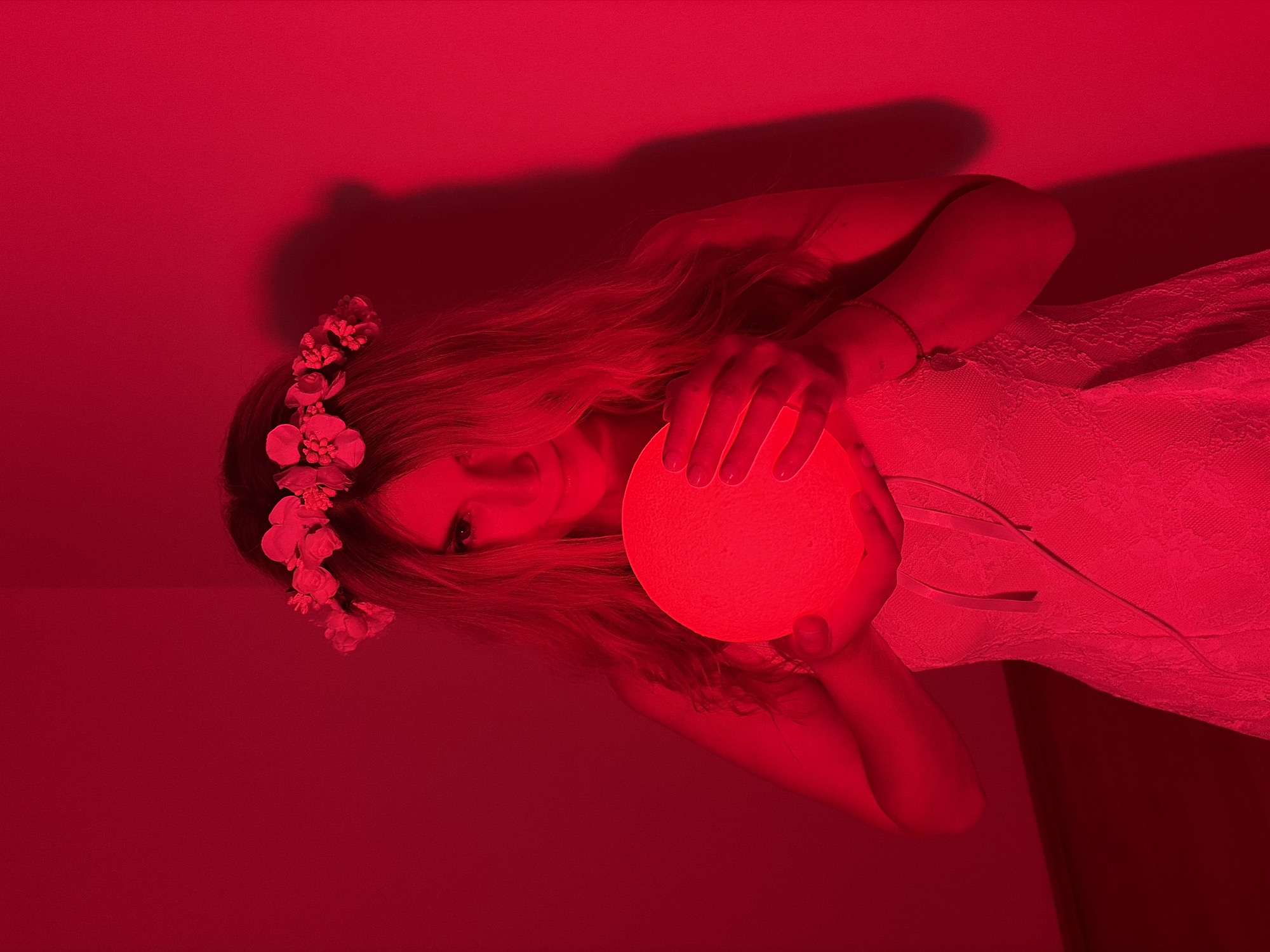 A young woman, illuminated by red light, is depicted indoors against a red wall. She is wearing a white lace dress and a flower crown on her head. She is holding a glowing red sphere in her hands, which casts light on her face. Her arms are crossed, and she is looking towards the viewer. The image is predominantly red due to the lighting. The city and its location in Budva, Montenegro are not discernible from the image.