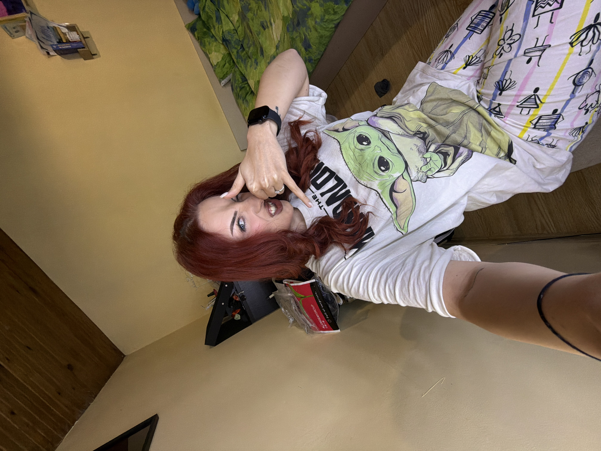 A selfie taken by a woman indoors, location unknown. The woman has red hair, wears a white t-shirt with a Baby Yoda graphic, and pajama pants with a cartoon design. She is making a "rock on" hand gesture and looking at the camera. In the background, there is a bed with a green patterned blanket, a black end table, and a package of wet wipes. The walls are painted a light beige color.