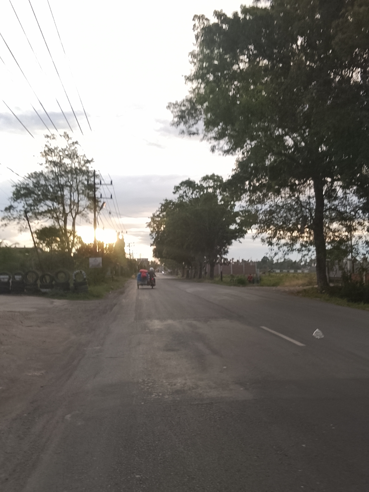 The image depicts a roadside scene in Meunasah Pupu, Indonesia, with a paved road stretching into the distance. The sun is low in the sky, casting a bright glow through the trees lining the left side of the road, creating lens flare. Several utility poles with visible wires are also present on the left. To the far left, a pile of tires is stacked against a grassy embankment. On the road, a three-wheeled vehicle, possibly a tricycle or auto-rickshaw, is traveling away from the viewer. The vehicle has a blue canopy and appears to have at least two occupants. The right side of the road is bordered by trees and a fence in the background. The sky is overcast with some clouds visible. The overall impression is a rural or semi-urban setting during the late afternoon or early evening.