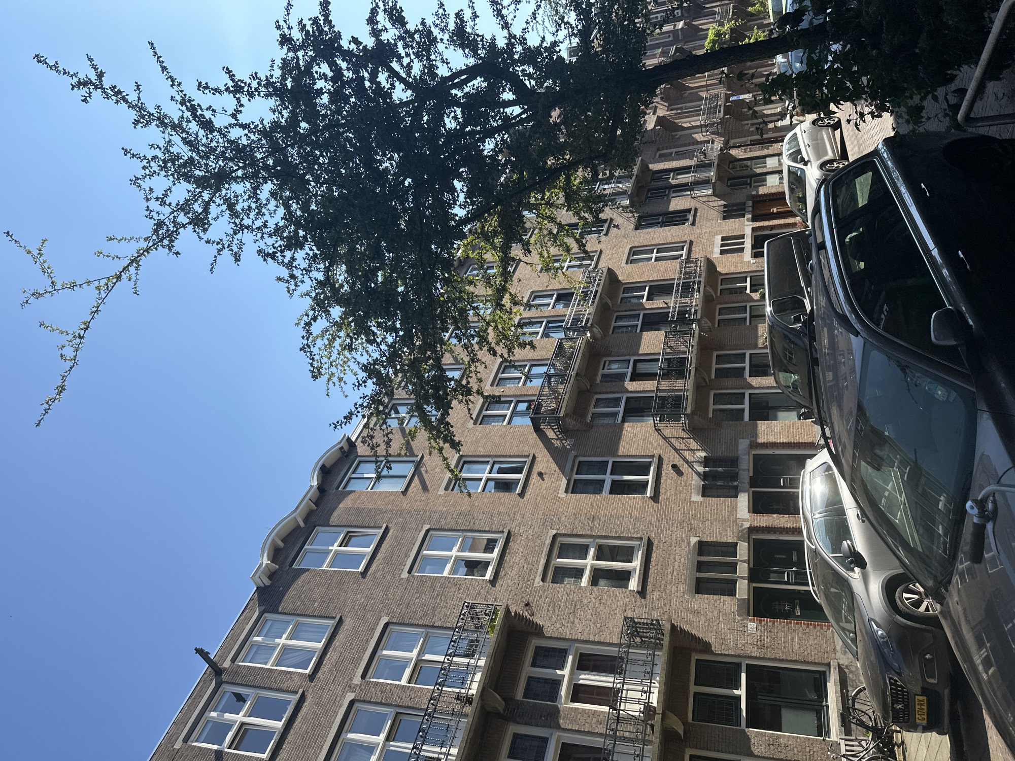 The image shows a street scene in Amsterdam, Netherlands. The foreground features several parked cars, with a dark-colored SUV closest to the viewer. Further down the street, a silver hatchback and a dark minivan are visible. The background consists of a multi-story brick building with numerous windows and balconies. A large tree with dark green foliage is positioned on the right side of the frame, casting shadows on the building and street. The sky is clear and blue, indicating a sunny day. The architecture of the building suggests it is residential, and the presence of parked cars and bicycles indicates an urban environment. The license plate on one of the cars reads "G-174-RK".