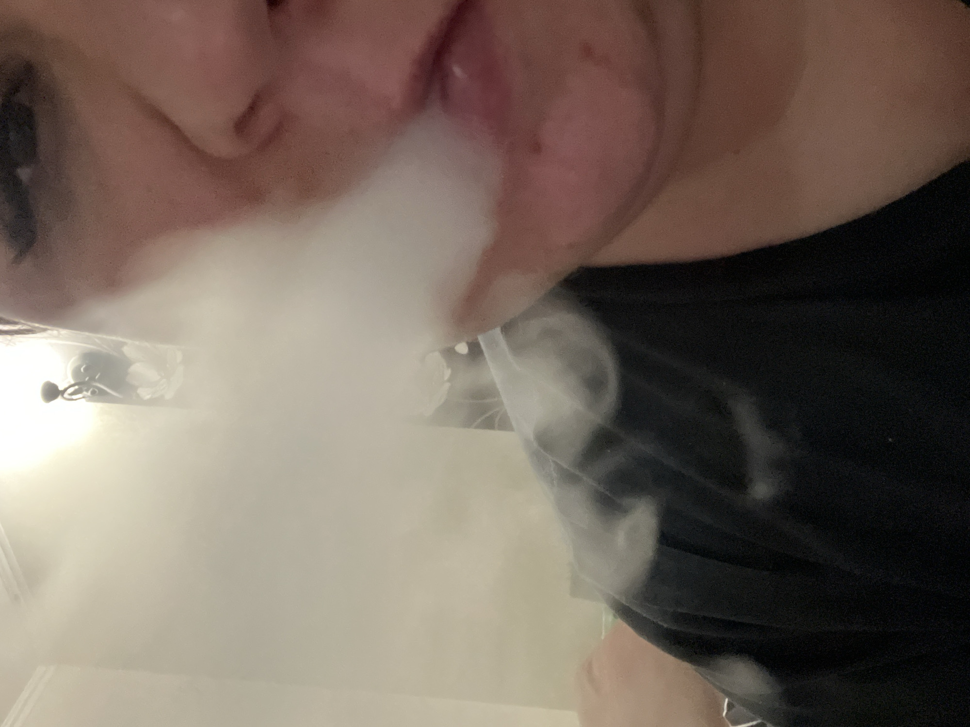 A close-up, low-angle shot focuses on a person exhaling a plume of white vapor. The person's face, including a visible eye, nose, and mouth, is positioned at the top of the frame. White smoke or vapor billows from the mouth, partially obscuring the view. The person is wearing a black t-shirt. In the blurred background, there is a ceiling fan and wall with a floral pattern. I do not have enough information to establish the location within the city of Chaikovsky, Russia.