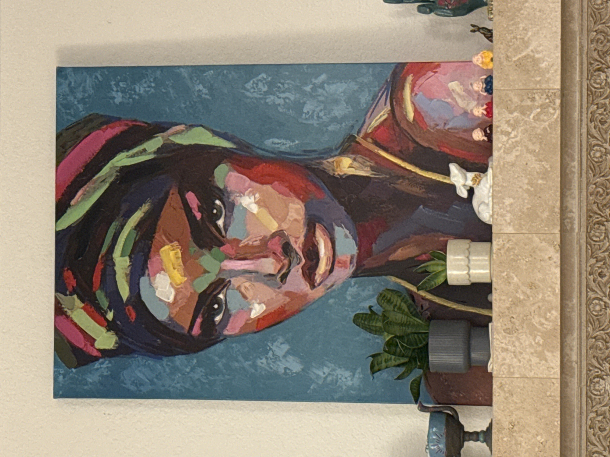 A painting of a woman with colorful brushstrokes is displayed on a light-colored wall, positioned above a marble fireplace mantel. The woman in the painting wears a vibrant headscarf and her face is rendered with a variety of colors, including pink, yellow, green, and blue. Her eyes are dark and piercing. The background of the painting is a textured teal color.
On the mantel, to the left of the painting, there is a small, dark blue chalice-like object on a pedestal. Next to it are two potted plants. The plant on the left is in a dark gray ribbed pot, and the plant on the right is in a small, white scalloped pot. To the right of the plants, a small white ceramic dog-like figure rests. Further to the right, there are five small figurines of what appear to be meditating figures, each wearing different colored robes. To the right of these figurines is a small black animal figurine, possibly a dog, and then a dark green decorative hand.
The fireplace mantel itself is made of light-colored, textured marble. Below the mantel, there is a decorative architectural element with an intricate carved pattern. The wall behind the painting and mantel is painted a light, neutral color. This scene is located within a residential interior.