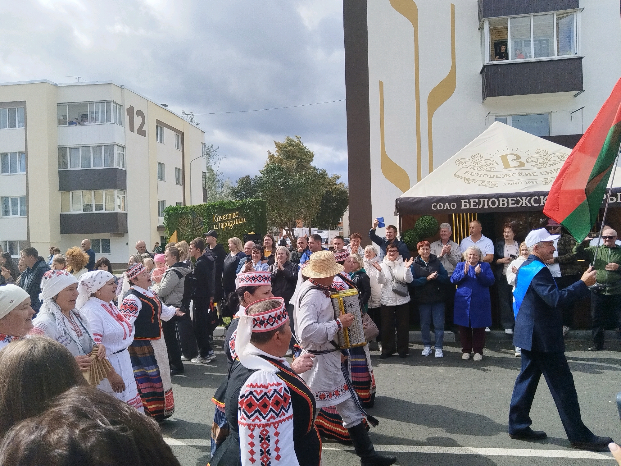 In a city square, a crowd gathers under an overcast sky, facing a building with beige and white vertical decorative lines. To the left, a multi-story residential building with balconies and the number "12" is visible. A white tent with "BELOVEZHSKIYE SYRY ANNO 1938" printed on it is set up in front of the building, partially obscuring a storefront. On the right, a person in a dark suit with a blue sash and white baseball cap walks forward, holding a green umbrella. A flag with red and green stripes is held by someone off-frame to the right. A group of people in traditional folk costumes, including women in white embroidered dresses and headscarves and men in embroidered shirts and vests, are in the foreground. One man plays an accordion. Other people in the crowd are watching, some clapping. The ground is paved.