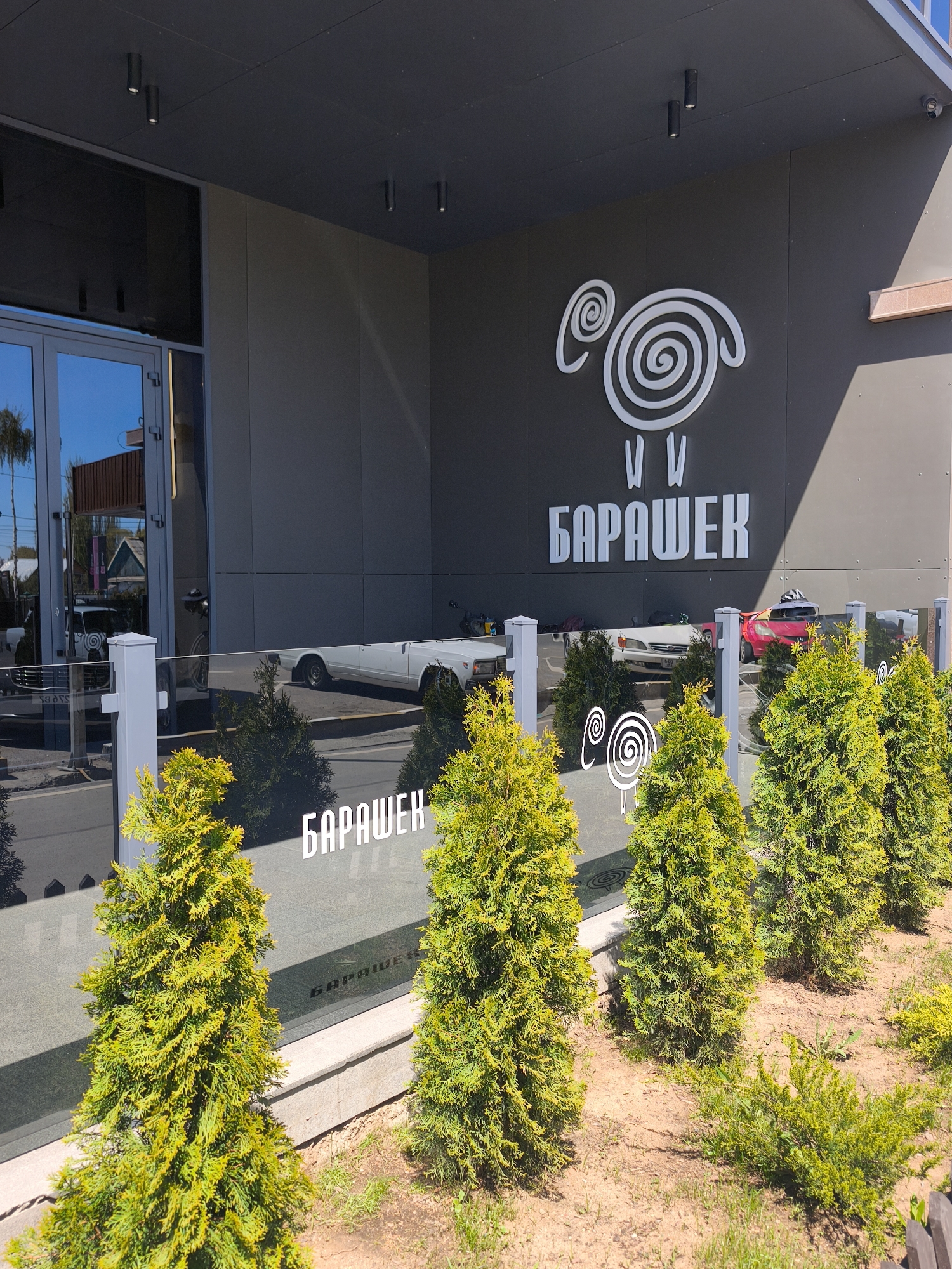 The image depicts the exterior of a building in Çolpon-Ata, Kyrgyzstan. A sign above the entrance shows a stylized image of a sheep with the word "БАРАШЕК," written in white Cyrillic letters, indicating that it could be a restaurant or shop. A glass barrier runs in front of the building, and reflections of parked cars and trees can be seen in the glass. Several small evergreen trees are planted in a row in front of the barrier on soil. The building has a modern architectural style with a gray façade.