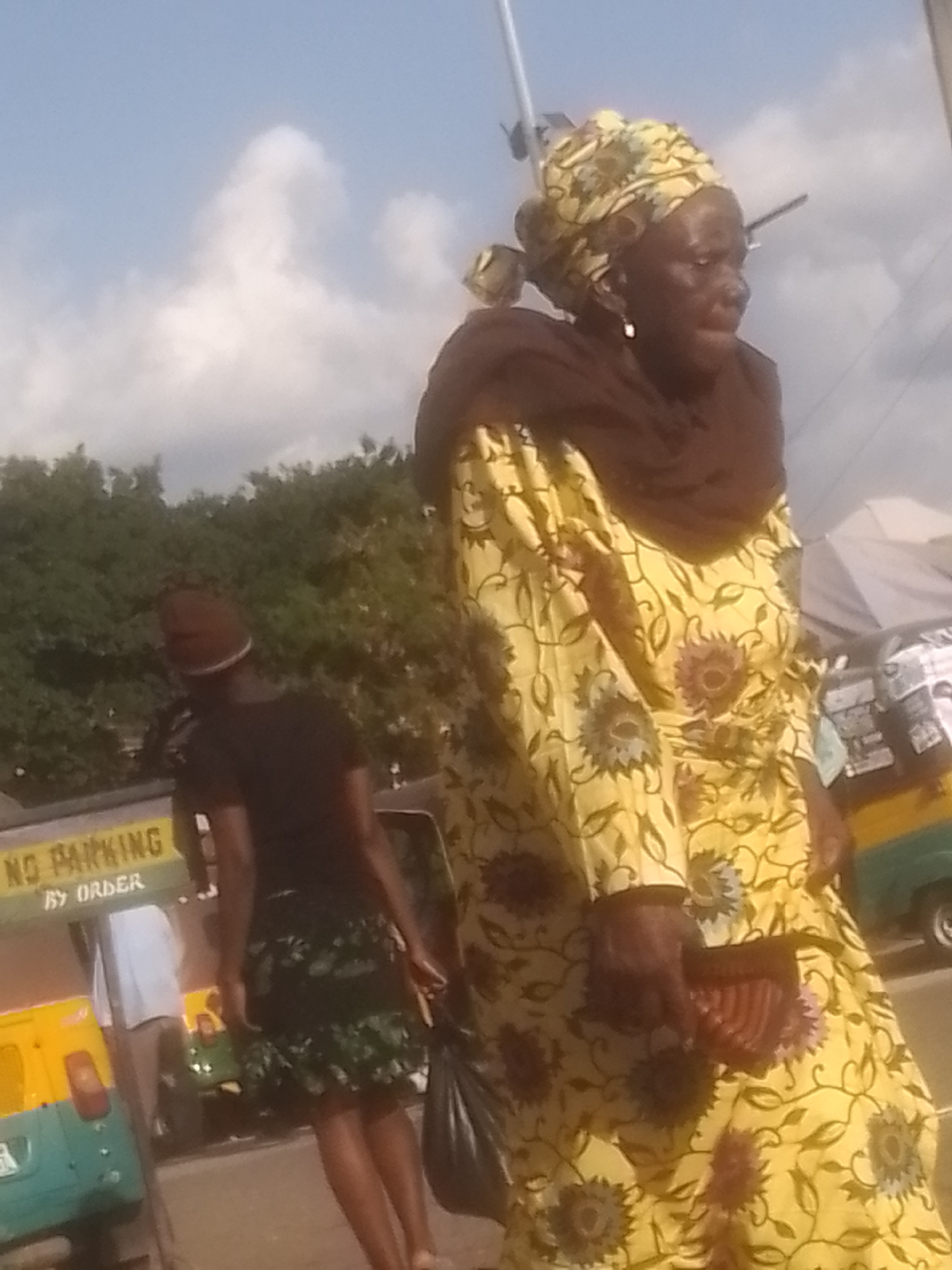 The image depicts an outdoor scene in the city of Gwong, Nigeria, under a bright, partly cloudy sky. In the foreground, a woman is prominently featured, wearing a vibrant yellow dress with a floral pattern and a brown headscarf. She appears to be walking, holding a dark-colored bag in her right hand and a pink object with a ridged surface in her left. To her left, another person is visible, wearing a dark t-shirt and a brown head covering, also appearing to be in motion. Behind these individuals, a yellow and green auto-rickshaw is parked. A yellow sign with the text "NO PARKING BY ORDER" is affixed to a pole in the background, partially obscured by the auto-rickshaw and the people. Trees and some structures can be seen further in the background.