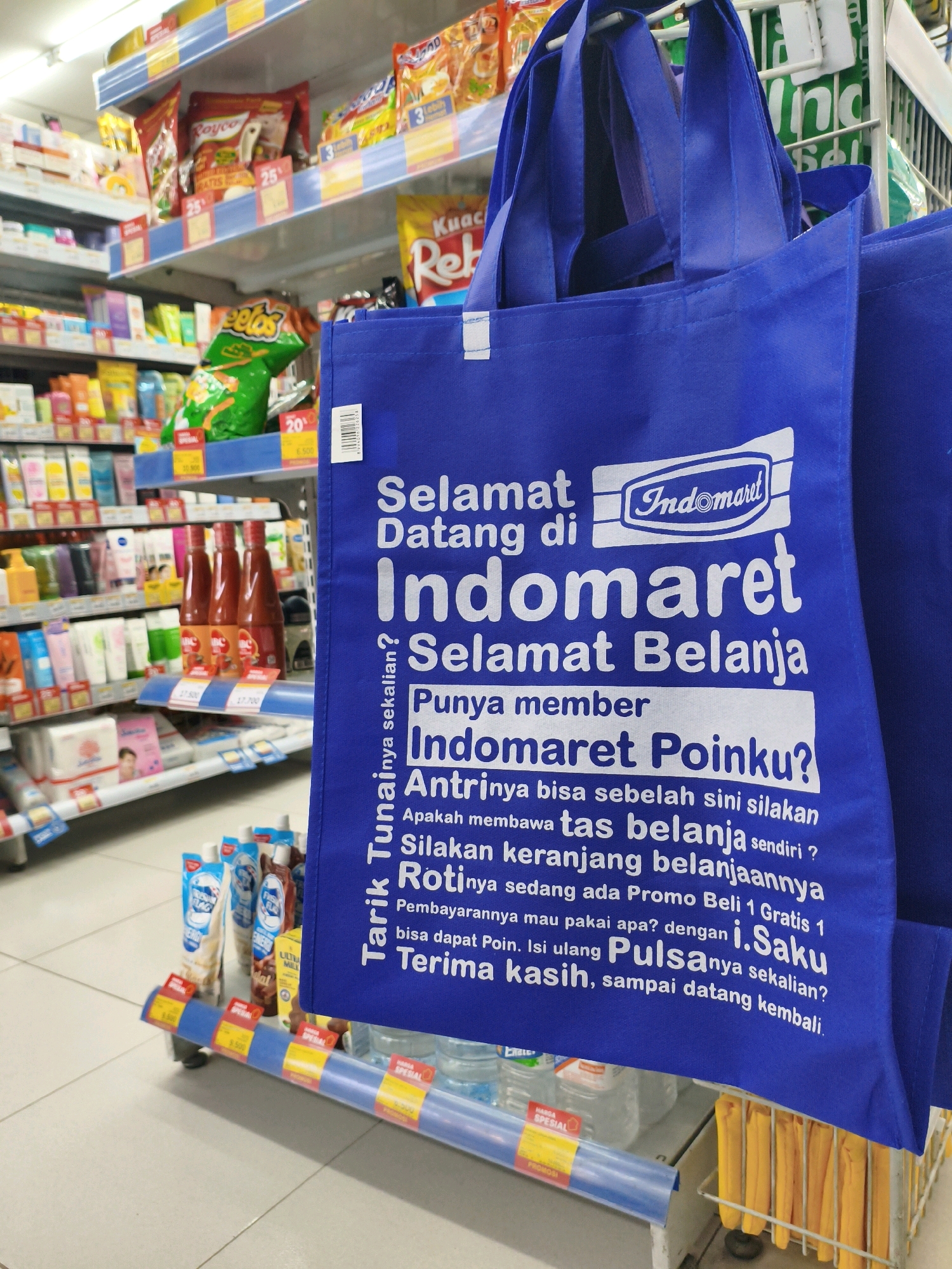 An eye-level, close-up shot captures a blue reusable shopping bag with Indonesian text and the Indomaret logo prominently displayed, hanging in front of shelves stocked with various products. The bag's text reads: "Selamat Datang di Indomaret Selamat Belanja. Punya member Indomaret Pointku? Antrinya bisa sebelah sini silakan membawa tas belanja sendiri? Silakan keranjang belanjaannya Roti nya sedang ada Promo Beli 1 Gratis 1 Pembayarannya mau pakai apa? dengan i.Saku bisa dapat Poin. Isi ulang Pulsa nya sekalian? Terima kasih, sampai datang kembali." The Indomaret logo features a stylized "i" within a rectangle and the text "Indomaret" in a script font. Behind the bag, shelves are filled with packaged snacks like Cheetos and Royco, as well as bottles of sauce and toiletries. The store environment appears to be a typical convenience store or minimarket. The presence of Indonesian text and the Indomaret branding indicates the location is likely in Indonesia, possibly within the city of Ngunut, a town in East Java, Indonesia, known for its Indomaret stores. The lighting is bright and evenly distributed, typical of an indoor retail setting.