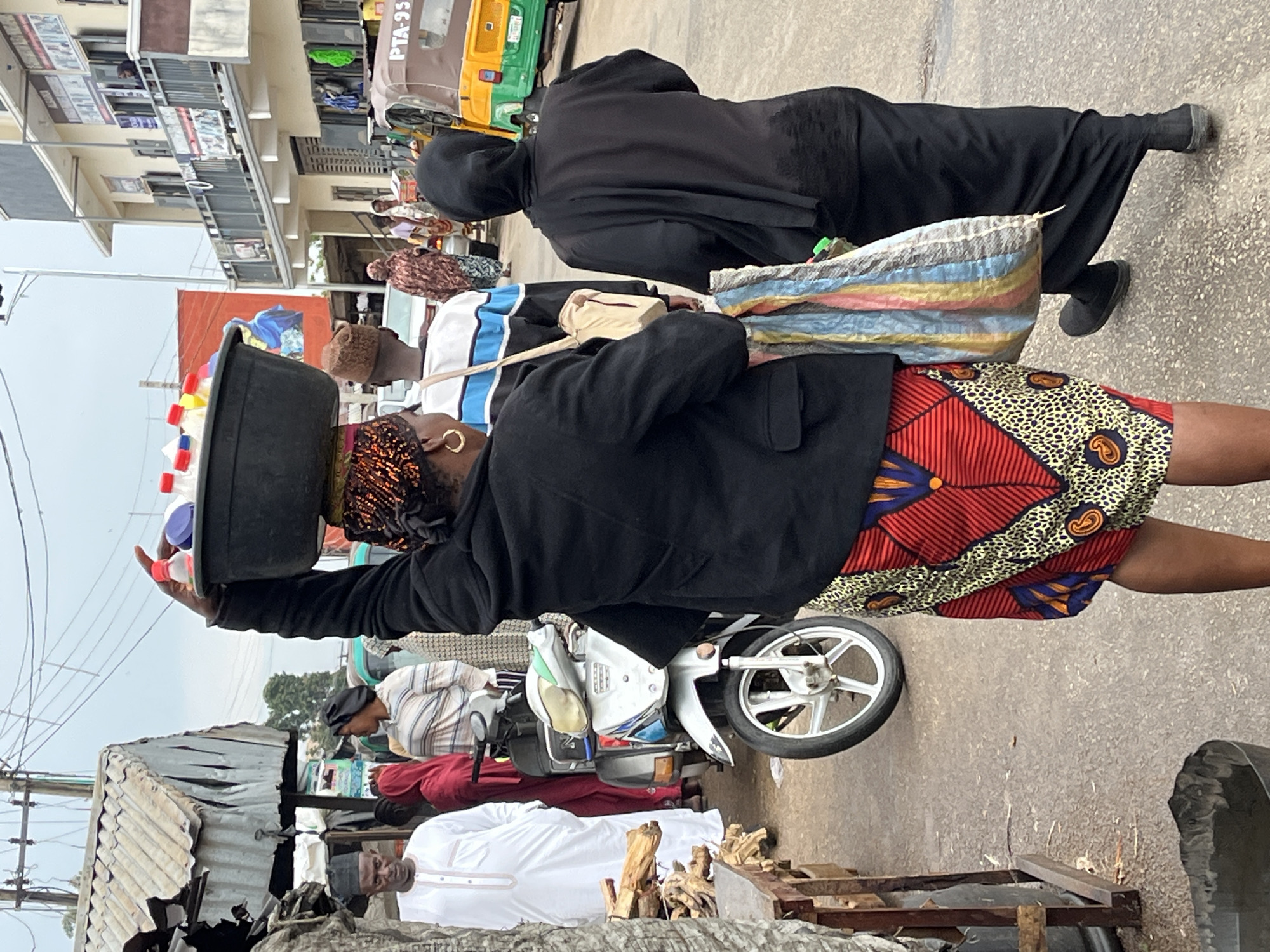 The image depicts a street scene in Jos, Nigeria. A woman in a black jacket and patterned red and yellow skirt balances a black basin filled with items (possibly containers) on her head as she walks on the pavement. Another woman clad in a black abaya follows closely behind. Behind them is a man in a white shirt with blue stripes. A motorcycle is parked nearby, as well as some people walking. The background includes a corrugated iron roof structure and buildings typical of a city setting, some featuring awnings. Further away, a "keke" or "tuk-tuk" type vehicle is visible.