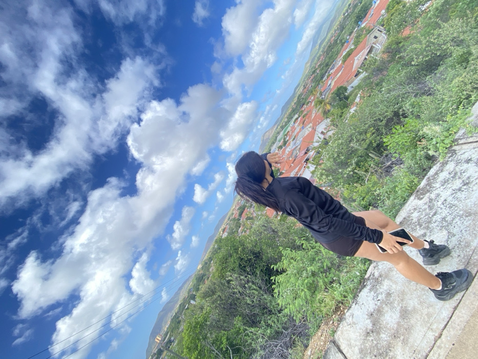 A young woman with dark hair in a ponytail, wearing a black long-sleeved shirt and shorts, stands on a concrete ledge, holding a phone. She shields her eyes, looking out over a landscape of green trees and orange-roofed buildings under a blue sky with fluffy white clouds. The view is from an elevated perspective, suggesting she's on a hill or mountain. The scene is angled.