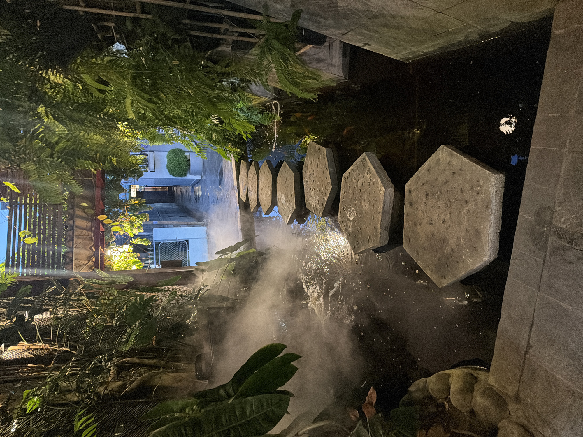 The image shows a pathway in Nha Trang Ward, Vietnam, illuminated at night. Hexagonal stepping stones traverse a narrow water feature. Mist rises from the water. Dense foliage frames the scene. Buildings are visible in the background.
