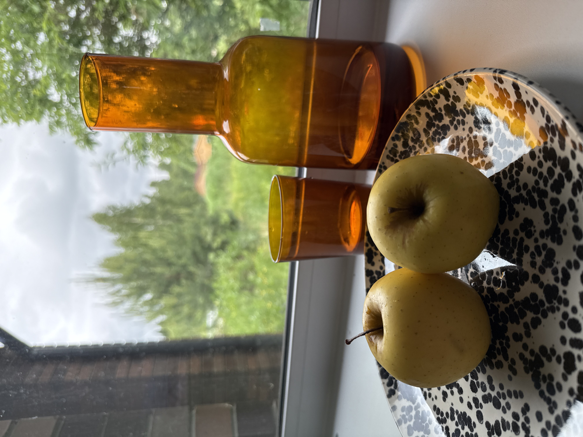 The image presents a still life arranged on a white window sill, with an outdoor scene visible through the window. In the foreground, two yellow apples, one retaining a short brown stem, are placed on a round plate. The plate features a high-contrast speckled pattern of irregular black shapes on a white background, with reflections of ambient light and some small water droplets visible on its glossy surface and on the apples. To the right of the plate, an amber-toned glass tumbler and a taller, similarly colored amber glass bottle or carafe stand on the sill.
The window pane itself shows minor smudges and dust particles, particularly on the left vertical edge of the visible window frame, which appears dark. Beyond the window, a lush, green natural environment is visible, characterized by numerous trees with dense foliage. The outdoor background is rendered with a soft, out-of-focus effect. The sky above is overcast, and a faint outline of a building with an orange or brown roof can be discerned through the trees in the mid-right portion of the background.
The image does not contain any specific visual cues, such as unique architecture, identifiable landmarks, or text, that would allow for the confirmation of its location as the city of Izhevsk, Russia. The depicted outdoor environment of trees and a distant structure is generic and could be found in various locations.
