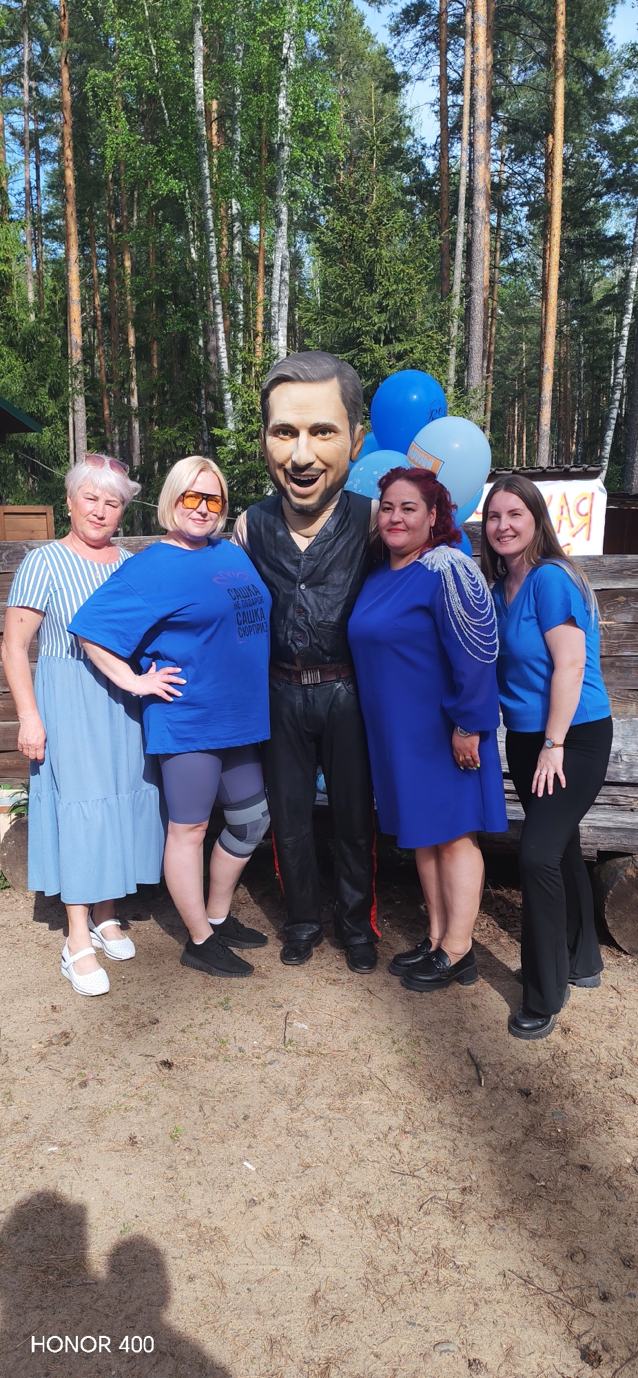 A group of five women poses with a life-size caricature statue of a man. The group stands on a sandy ground in an outdoor setting, possibly a park or recreational area, as suggested by the forest backdrop with trees. The woman furthest to the left wears a striped blue and white dress. The woman next to her wears a blue t-shirt and shorts, with a knee brace on her left knee. The statue depicts a man in a dark leather vest and pants. The woman next to the statue is wearing a blue dress. The woman on the right is wearing a blue shirt and black pants. Blue balloons and a sign with Cyrillic writing are visible behind the group. The camera's mark, "HONOR 400" is located on the lower portion of the frame.