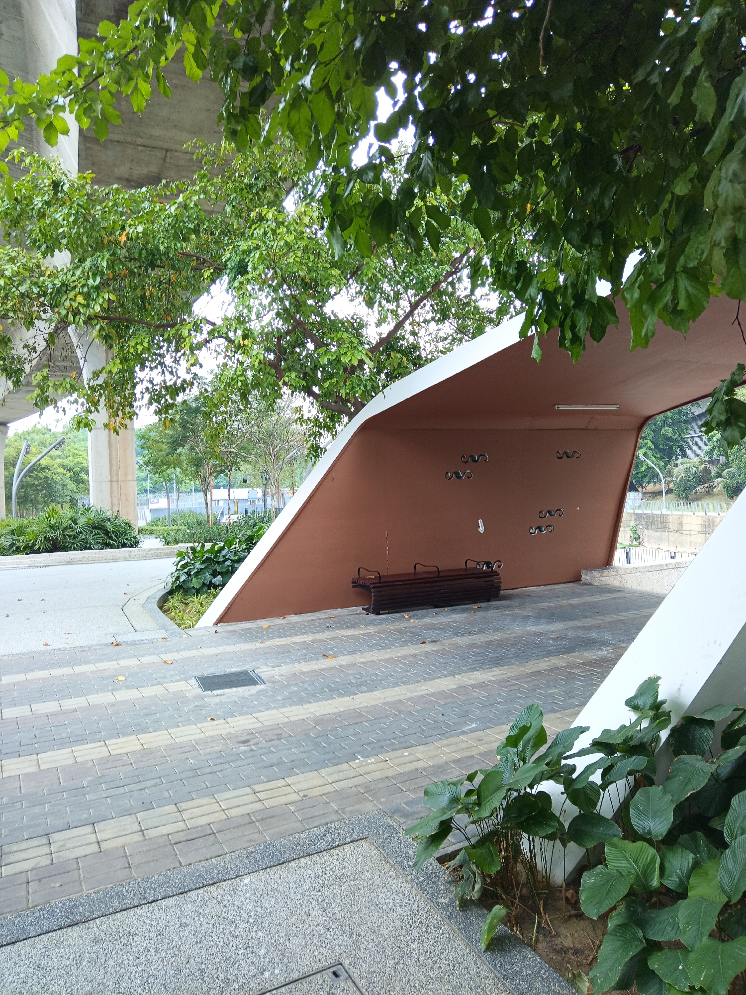 The image depicts an outdoor scene in Kuala Lumpur, Malaysia, under a large concrete overpass. In the foreground, there is a walkway paved with interlocking bricks in alternating shades of grey and beige. To the right, a modern, curved shelter with a brown interior and white exterior panels is visible. Inside the shelter, a wooden bench is placed on the paved floor. The brown wall of the shelter features several decorative, wavy black elements. Lush green plants grow in a planter box to the right of the shelter, adjacent to the walkway. In the background, trees and some low-rise buildings can be seen, with more trees lining the horizon. The overall impression is of a public space designed with contemporary architecture integrated with natural elements, likely a bus stop or waiting area beneath a busy road.