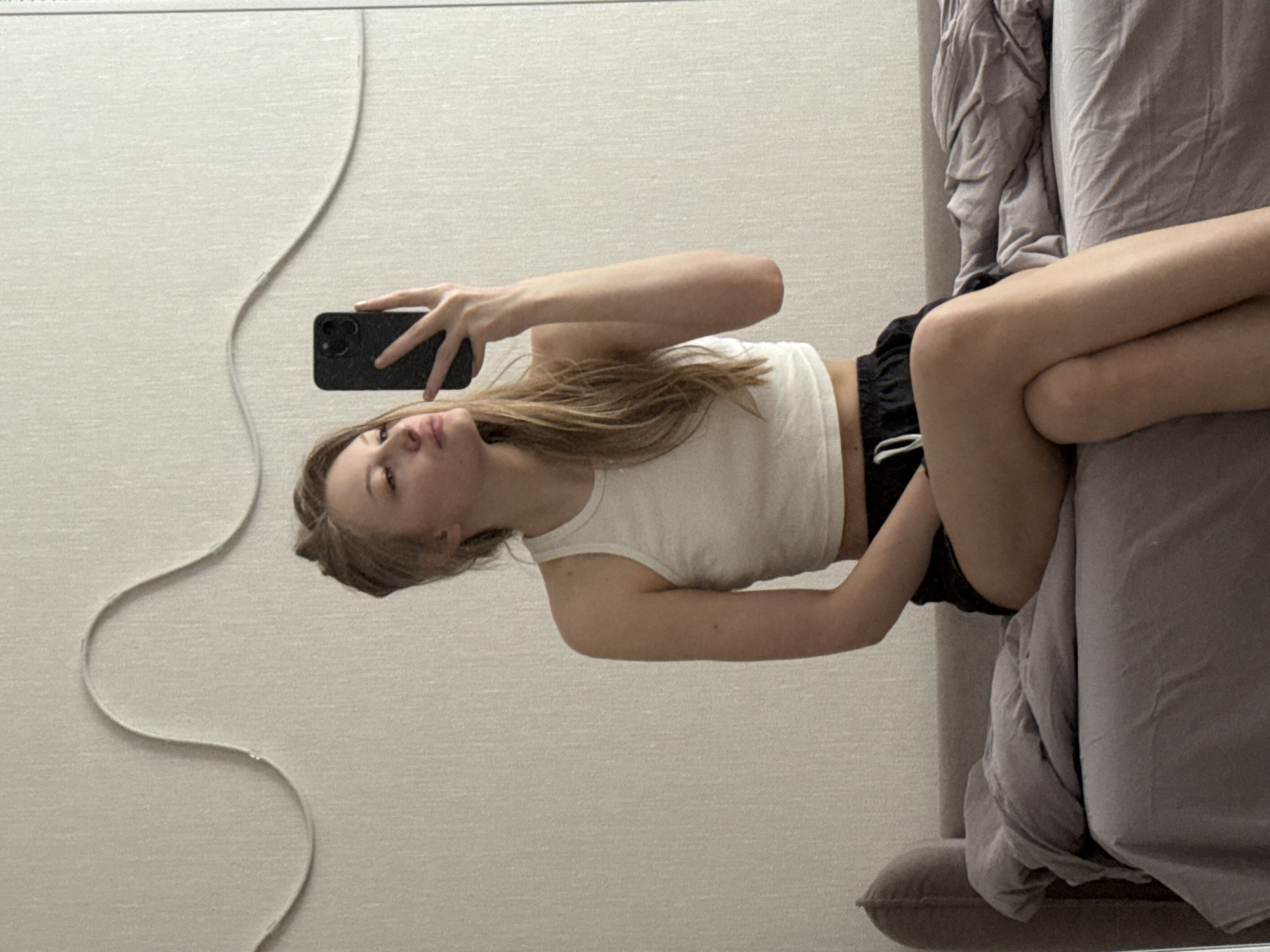 A young woman is visible in a mirror selfie. She is wearing a white tank top and black shorts, lying on a bed. The background shows a wall and part of the bed with a gray blanket. The location is not definitively discernible in this image, making it impossible to confirm the claim of Perm, Russia. The image does not contain any signs or symbols that could be used to determine the city.