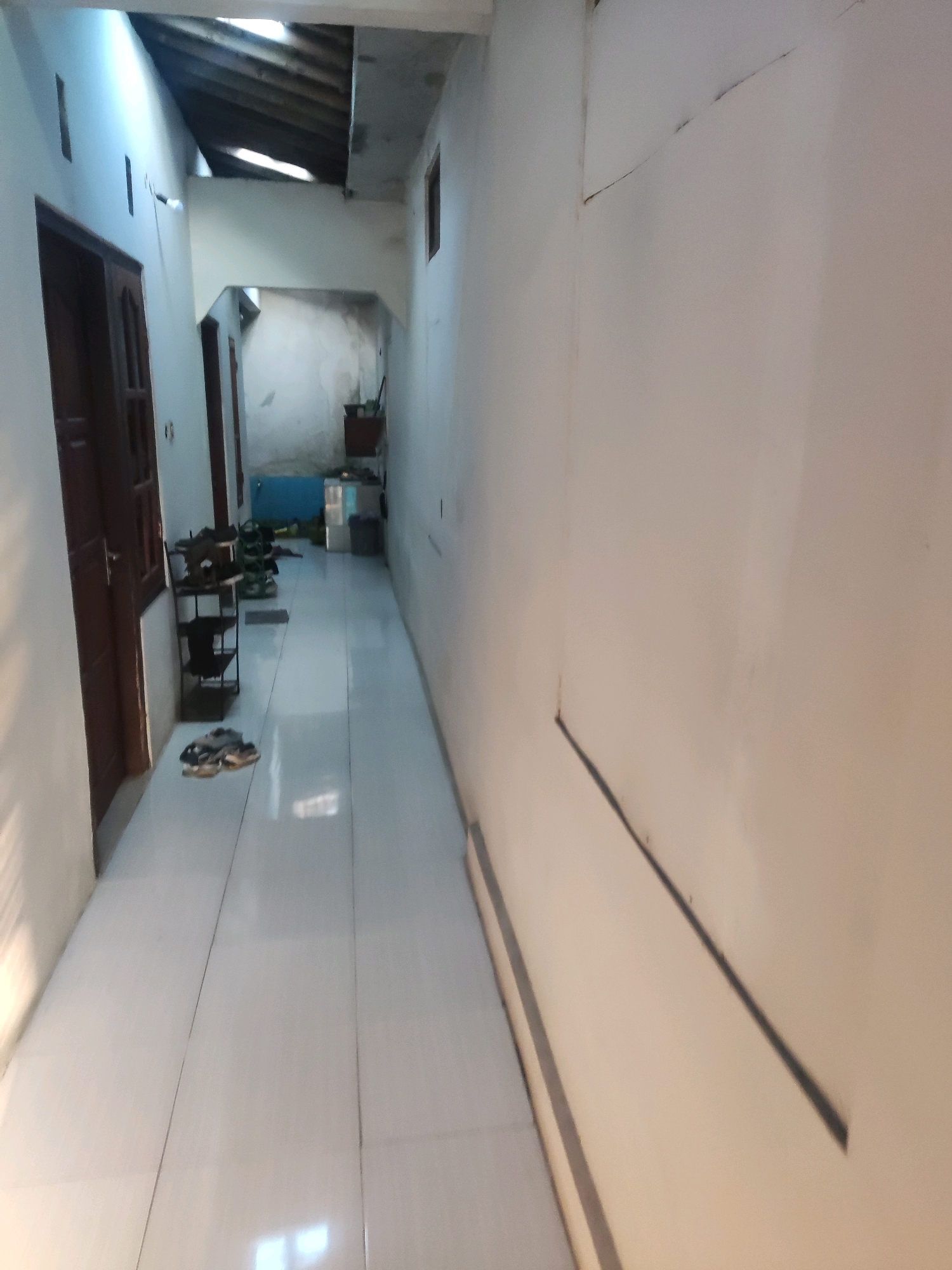 The image depicts a long, narrow, indoor hallway or corridor with white tiled floors and white walls. The hallway appears to be part of a residential property, possibly a home or a guesthouse, located in Trihanggo, Indonesia.
On the left side of the hallway, there is a dark brown wooden door. Further down the hallway on the left, a black metal shoe rack is visible, holding several pairs of shoes and sandals. Beyond the shoe rack, another door is present.
The ceiling of the hallway shows exposed wooden beams and some sections of unfinished construction, suggesting the property may be under renovation or has an open architectural style. Light enters the hallway from an opening in the ceiling, creating bright patches on the floor.
On the right side of the hallway, the wall is plain white and appears to have some faint dark lines or cracks. At the far end of the hallway, there is a glimpse of what might be a kitchen or storage area, with cabinets and a blue object.
The overall impression is of a simple, utilitarian space. There are no people visible in the image. The scene is static and well-lit by artificial and natural light.