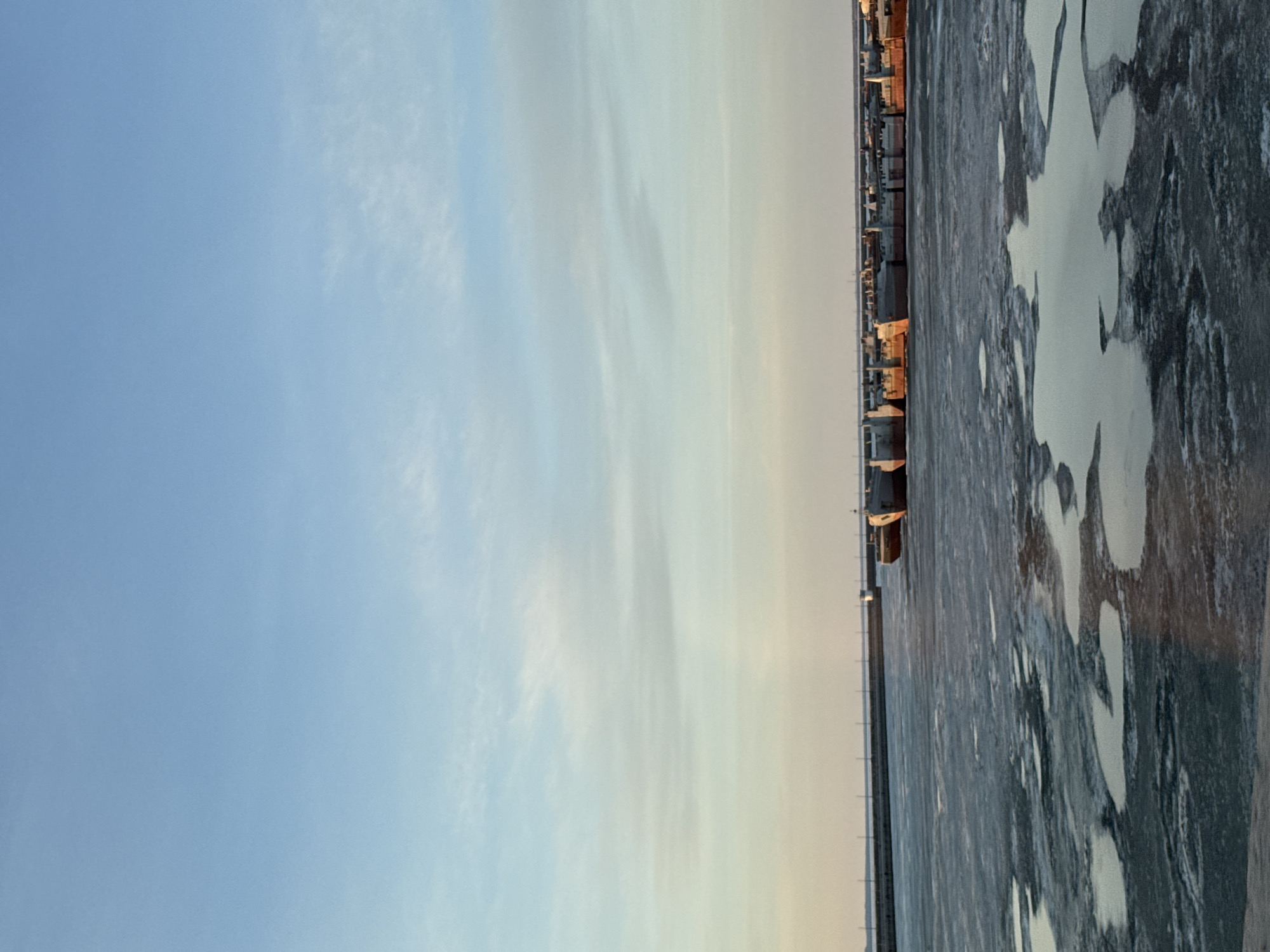 This media file captures a wide, serene landscape, likely taken during either sunrise or sunset given the warm, low-angle light illuminating the distant horizon. The image is divided into a dominant sky and a lower section of water and ice.
The sky occupies the upper two-thirds of the frame, displaying a soft gradient. On the left, it's a clear, light blue with faint, wispy clouds. Towards the right horizon, the sky shifts to a hazy, gentle golden-yellow, suggesting the sun's position.
Along the distant horizon line, a narrow strip of land is visible, populated by indistinct structures that appear to be a mix of low-rise buildings, possibly industrial or residential, catching the warm, golden light.
The lower portion of the image is a vast expanse of water, significantly covered in ice. Large, irregular patches of white and lighter grey ice stand out against darker areas, which may be open water or thinner ice. The texture of the ice indicates a cold environment.
The scene is devoid of any visible people or animals, contributing to a sense of tranquil vastness. The presence of extensive ice suggests a winter or early spring setting, consistent with the location context of Chaikovsky, Russia, likely depicting a section of the Votkinsk Reservoir. No visible text is present in the image.