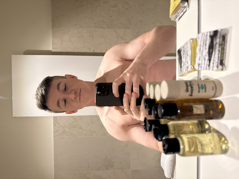 A shirtless male, approximately in his twenties to thirties, is depicted in a mirror selfie, holding a black smartphone with both hands at chest level. His head is slightly tilted down, and his eyes are looking towards the phone, with a slight, neutral expression on his face. The man has short, dark brown hair. The background shows a modern bathroom environment. A large, rectangular mirror occupies most of the upper frame. Below the mirror, a white countertop is visible, upon which several hotel-style amenities are arranged. These include multiple small bottles of liquid products (some clear, some amber) with black or white caps, and several individually wrapped bars of soap. One white bottle distinctly reads "Green Beauty" and "THE MERCHANT OF VENICE." Adjacent to these, two bars of soap in paper wrappers, patterned with black and white tree bark, are visible. The walls feature light-colored, possibly marble or tiled, surfaces. The presence of toiletries from "THE MERCHANT OF VENICE" brand indicates the scene is located in a hotel bathroom in the city of Venice, Italy.