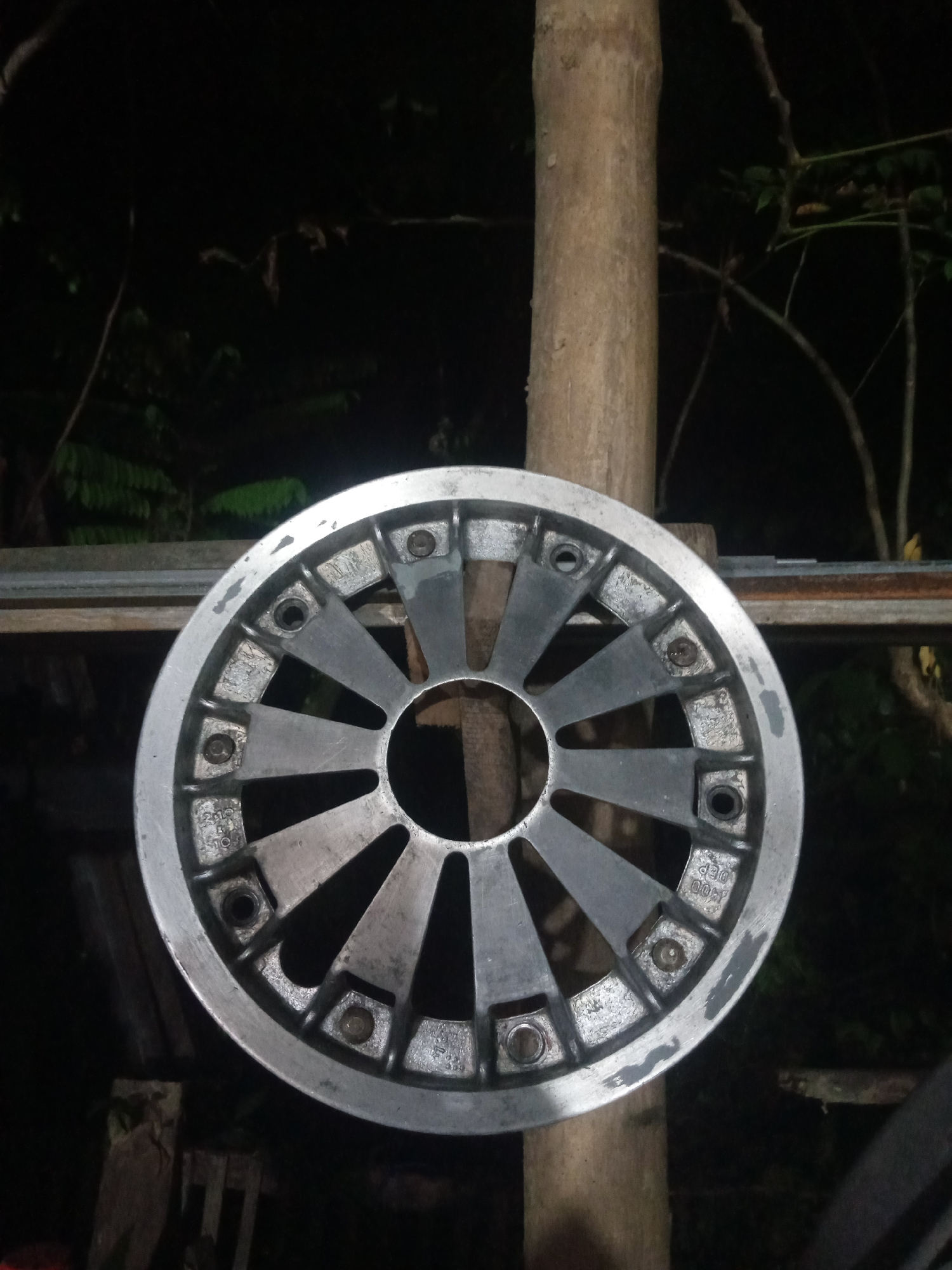 A silver, multi-spoke rim is mounted on a rough, weathered wooden post. The rim appears to be made of metal and has visible bolts and what look like bolt holes around its circumference. The spokes are angular and fan out from the center. The background is dark and out of focus, with glimpses of foliage and other wooden structures, suggesting an outdoor, possibly cluttered or storage area. The overall impression is of an object displayed or stored in a rustic setting. This scene is located in Balee Rastong, Indonesia.