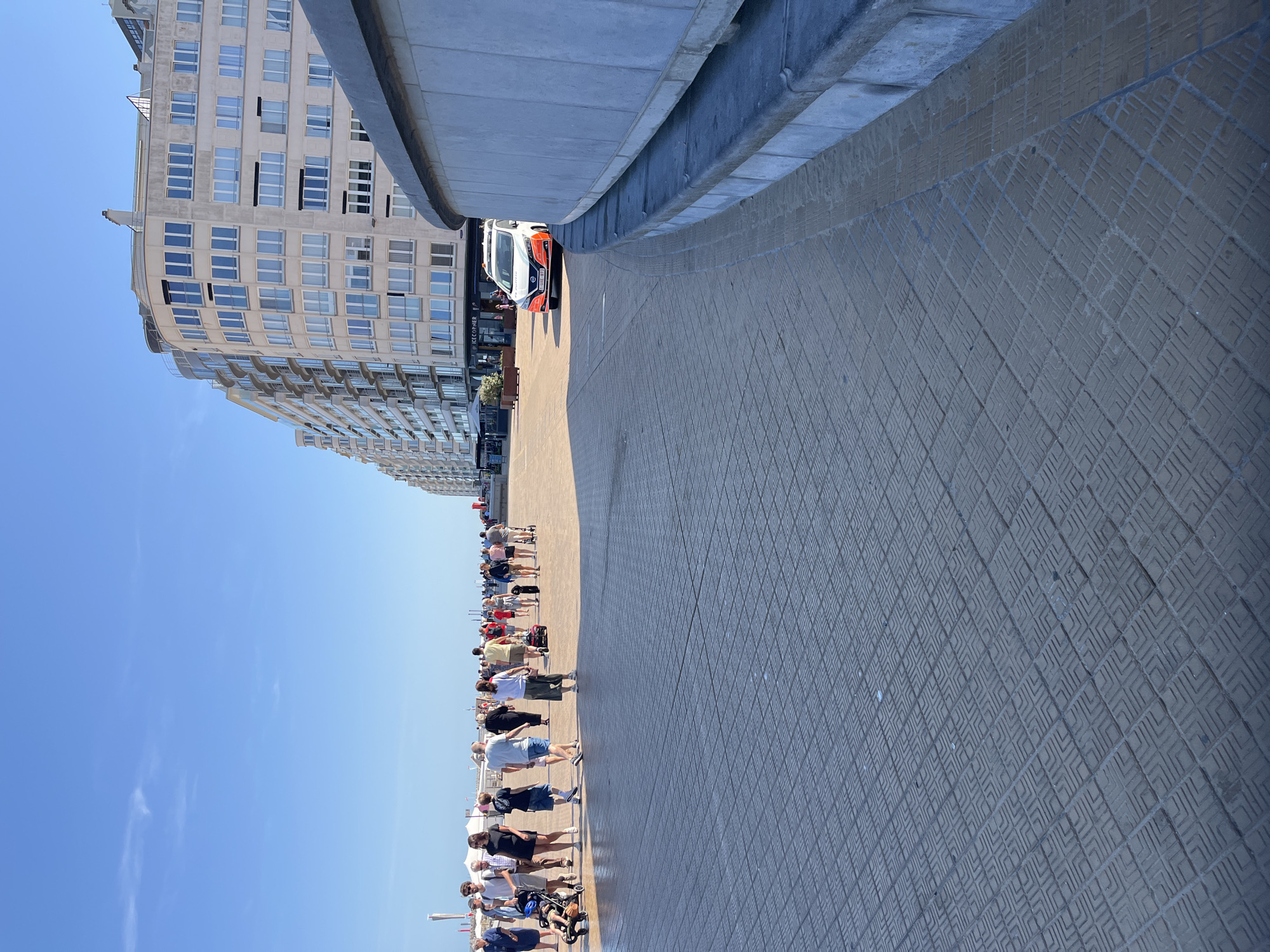 This is a vertical, eye-level, medium shot of a promenade in Ostend, Belgium, on a clear, sunny day. The foreground is dominated by a patterned pavement made of dark gray interlocking tiles. To the right, a curved, light gray concrete wall with a blue-gray stone coping separates the promenade from an unseen lower area. In the middle ground, a diverse crowd of people strolls along the promenade, some walking in groups, others individually. A man pushes a stroller, and a woman walks with a child. A police car, marked with "Politie" and a Belgian license plate, is parked on the road to the left of the building. The background features a long, curved, multi-story building with numerous windows, appearing to be apartment or hotel units, painted a light beige. Further back, the sky is a vivid, cloudless blue. The overall scene depicts a lively public space on a pleasant day in Ostend.