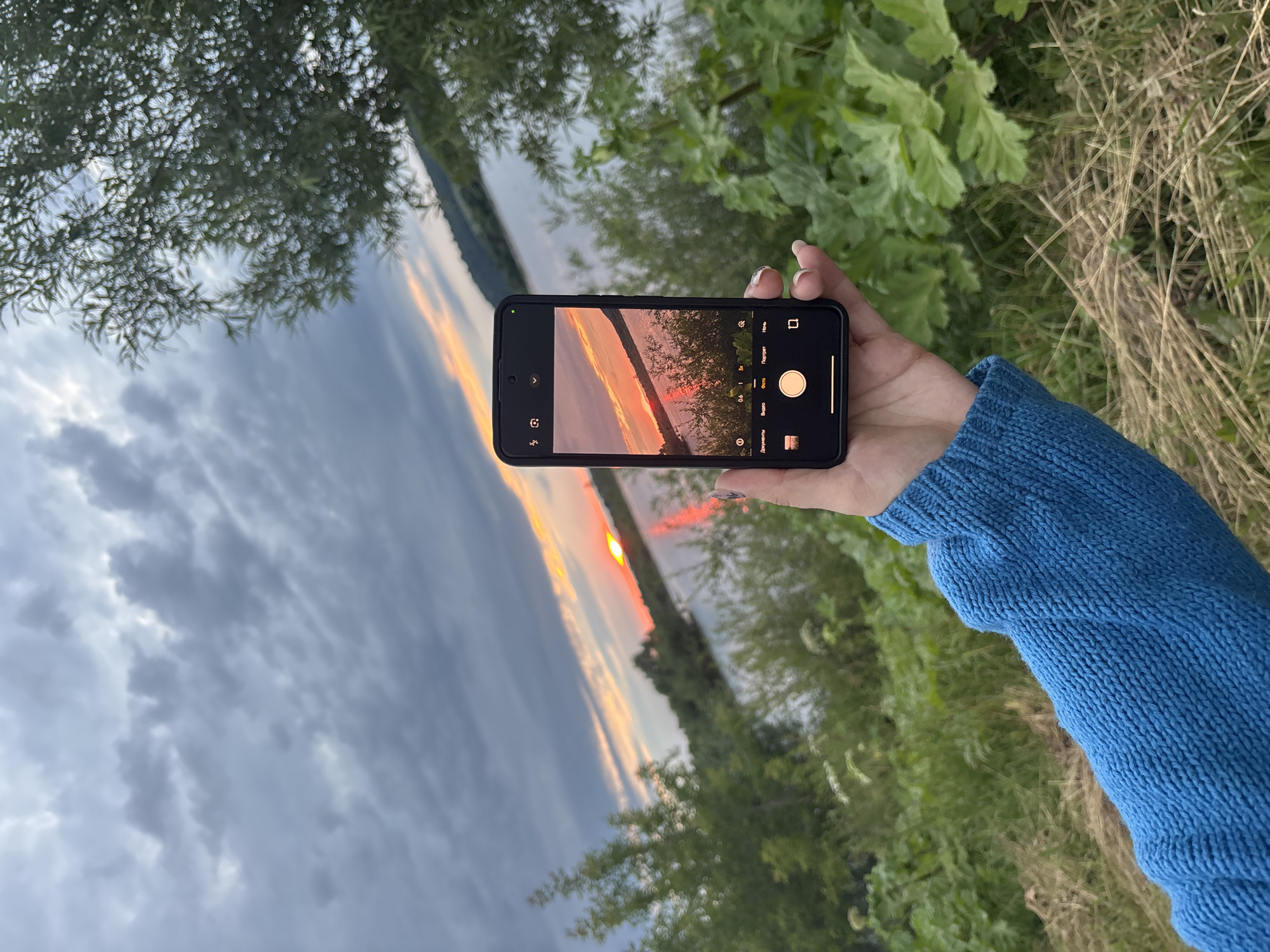 A hand wearing a blue knitted sweater holds a smartphone, capturing a sunset over a body of water. The phone's screen displays a camera interface with various shooting modes, including "Portret" (Portrait), "Foto" (Photo), and "Video" (Video). The image on the phone shows the sun setting on the horizon, casting orange and red hues across the sky and reflecting on the water. Trees with green foliage line the opposite shore, and dark clouds are visible in the upper left portion of the scene. The overall setting appears to be outdoors, near a river or lake, with lush greenery and dry grass in the foreground. The time of day is clearly sunset, indicated by the warm colors in the sky.