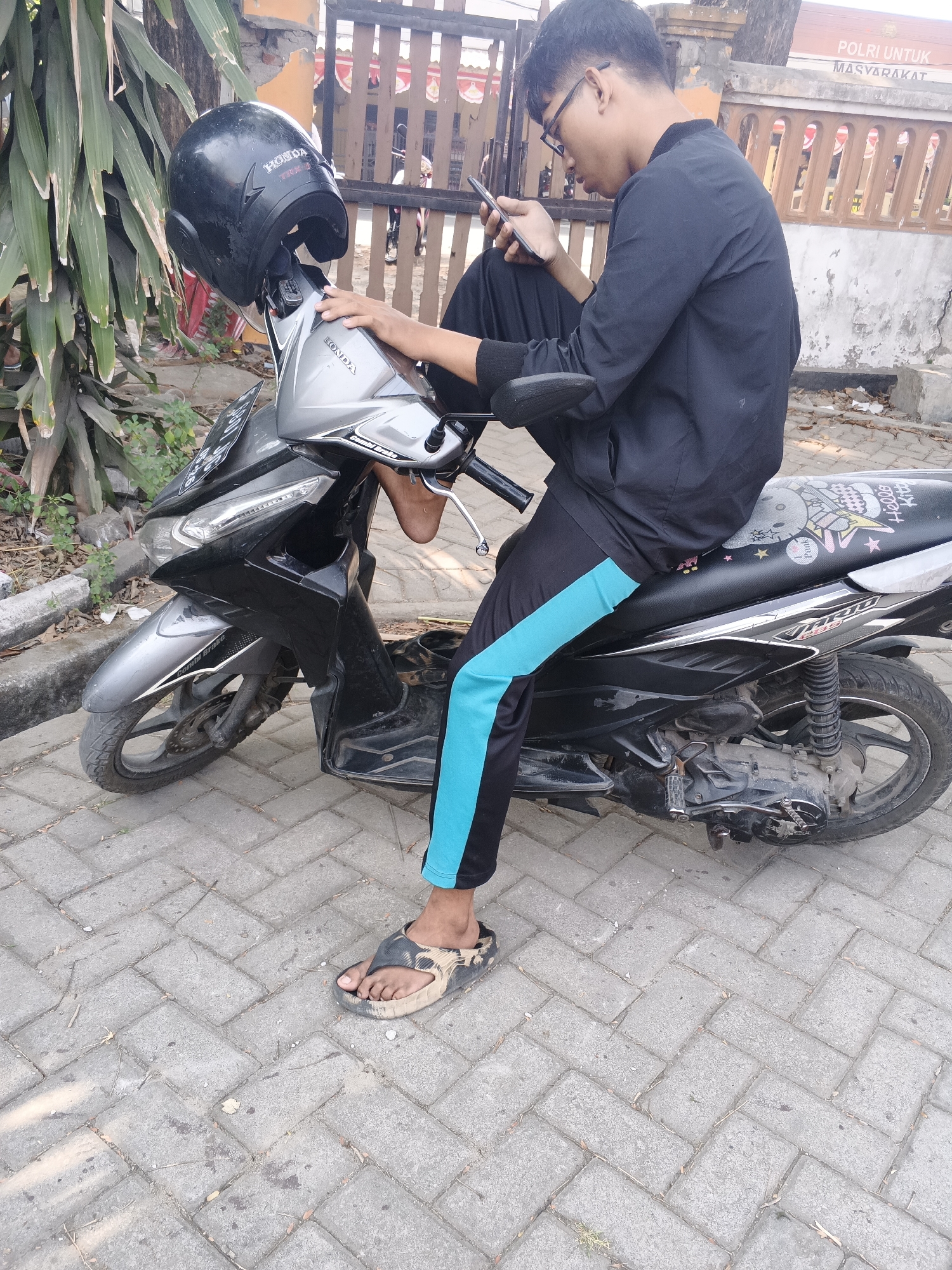 A young man wearing glasses, a black jacket, and black pants with a turquoise stripe on the side, sits on a black scooter. He is holding a mobile phone in his right hand and looking at it. A black helmet with a "HONDA" logo is placed on the front of the scooter. The scooter is parked on a paved sidewalk in Krian, Indonesia. In the background, there is a wooden fence and a building with a sign that reads "POLRI UNTUK MASYARAKAT" (Police for the People). The environment is outdoors, with some greenery and fallen leaves on the ground.