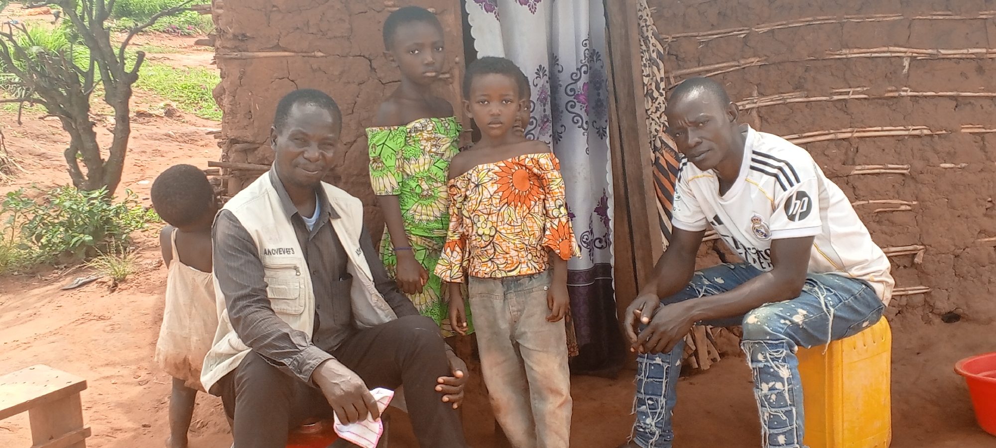 The image captures a group of six people outside a simple, earthen-built structure in what appears to be the city of Makungu, Democratic Republic of the Congo. On the left, a man wearing a light-colored vest with lettering that includes "ANOVEVE" and dark trousers sits on a small stool and holds a folded cloth. To his left, a small child in a light-colored dress stands facing away from the camera. Directly behind him are two young girls, dressed in off-the-shoulder tops with bright floral patterns, one green and one orange. To the right, a man wearing a white Real Madrid soccer jersey and patterned jeans sits on a yellow plastic container. A curtain hangs behind the two girls. The earthen structure behind the group appears to be a house, and the surrounding environment is dusty with visible trees and vegetation. A red plastic basin sits next to the man on the right.