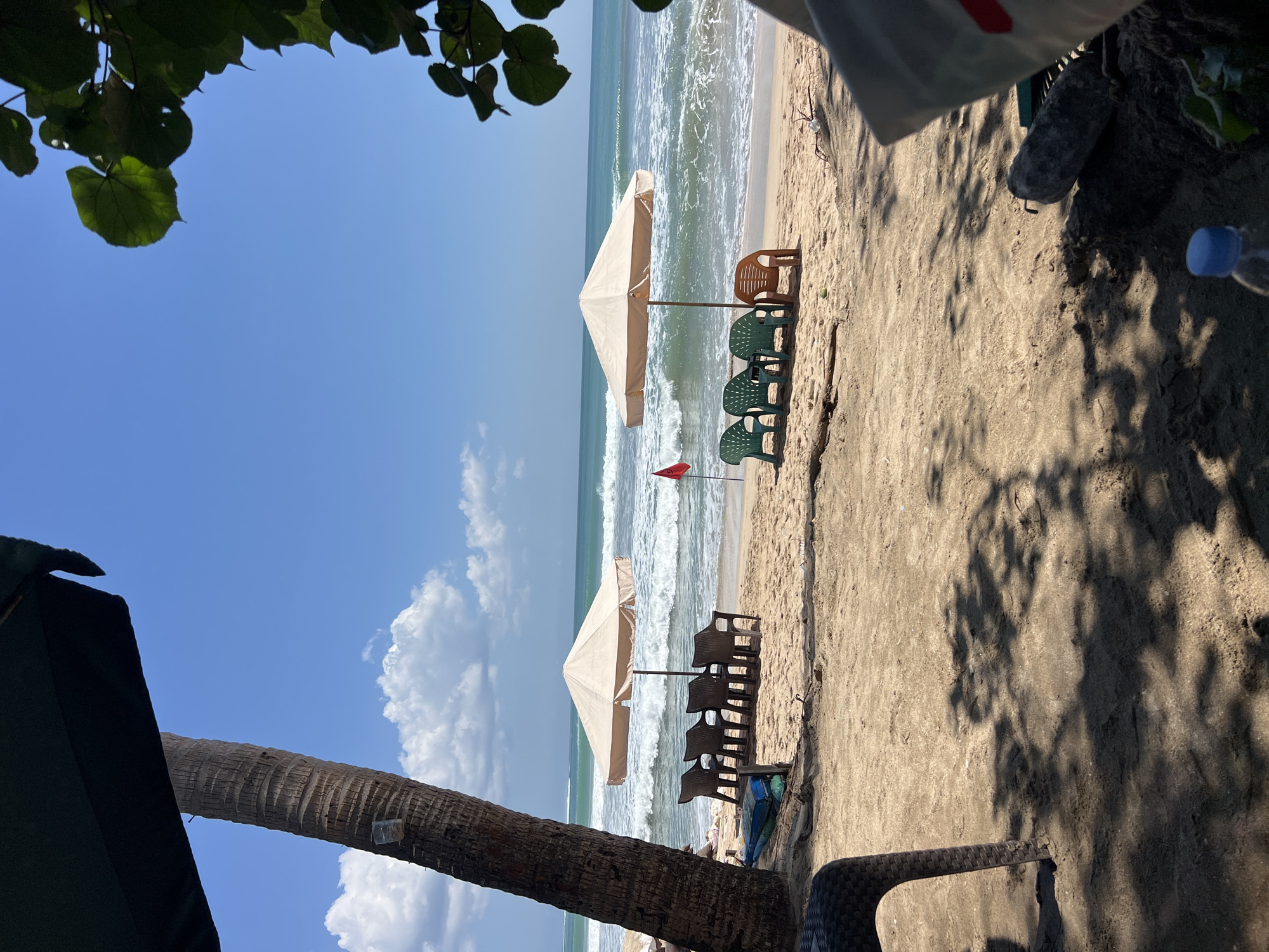 A serene view of a beach in Kuta, Indonesia, unfolds under a clear blue sky dotted with fluffy white clouds. The foreground is dominated by the sandy beach, casting dappled shadows from unseen foliage overhead. To the left, the rough texture of a palm tree trunk rises, partially obscured by a dark green umbrella.
Further back, two beige beach umbrellas stand sentinel over rows of plastic chairs, creating inviting spots for relaxation. The left umbrella is surrounded by four dark plastic chairs arranged in a line. To the right, a similar umbrella is accompanied by three green plastic chairs and one orange plastic chair, all facing the ocean. A small red flag planted in the sand adds a splash of color near the water's edge.
The ocean, with its gentle waves, stretches to the horizon, its turquoise surface glinting under the sun. The sandy shore meets the water, a clear demarcation between land and sea. In the immediate foreground, a black plastic chair is visible on the left, and on the right, a large rock and part of a bag are present. A plastic water bottle with a blue cap sits in the lower right corner. The overall atmosphere is one of peaceful tranquility, a perfect beach day in Kuta.