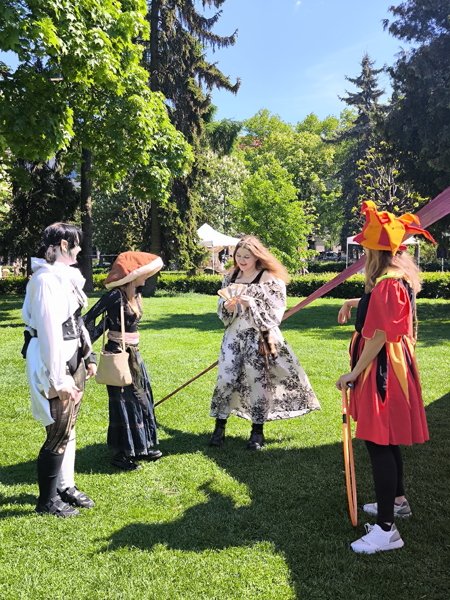 Four individuals dressed in costumes stand on a grassy area in a park. The person on the left wears a white tunic with black details, knee-high socks, and black shoes. Next to them, another person has a wide-brimmed brown hat, a dark-colored top, a long skirt, and a beige purse. The individual in the center wears a white dress with black floral patterns and is holding a fan of cards. On the right, a person is wearing a red and yellow jester's hat and a red dress with a black underlayer, leggings, and white sneakers, holding a long, orange object. The setting is a park with trees, a tent in the background, and a structure that can be seen as a building. The location is Riga, Latvia, according to the prompt.