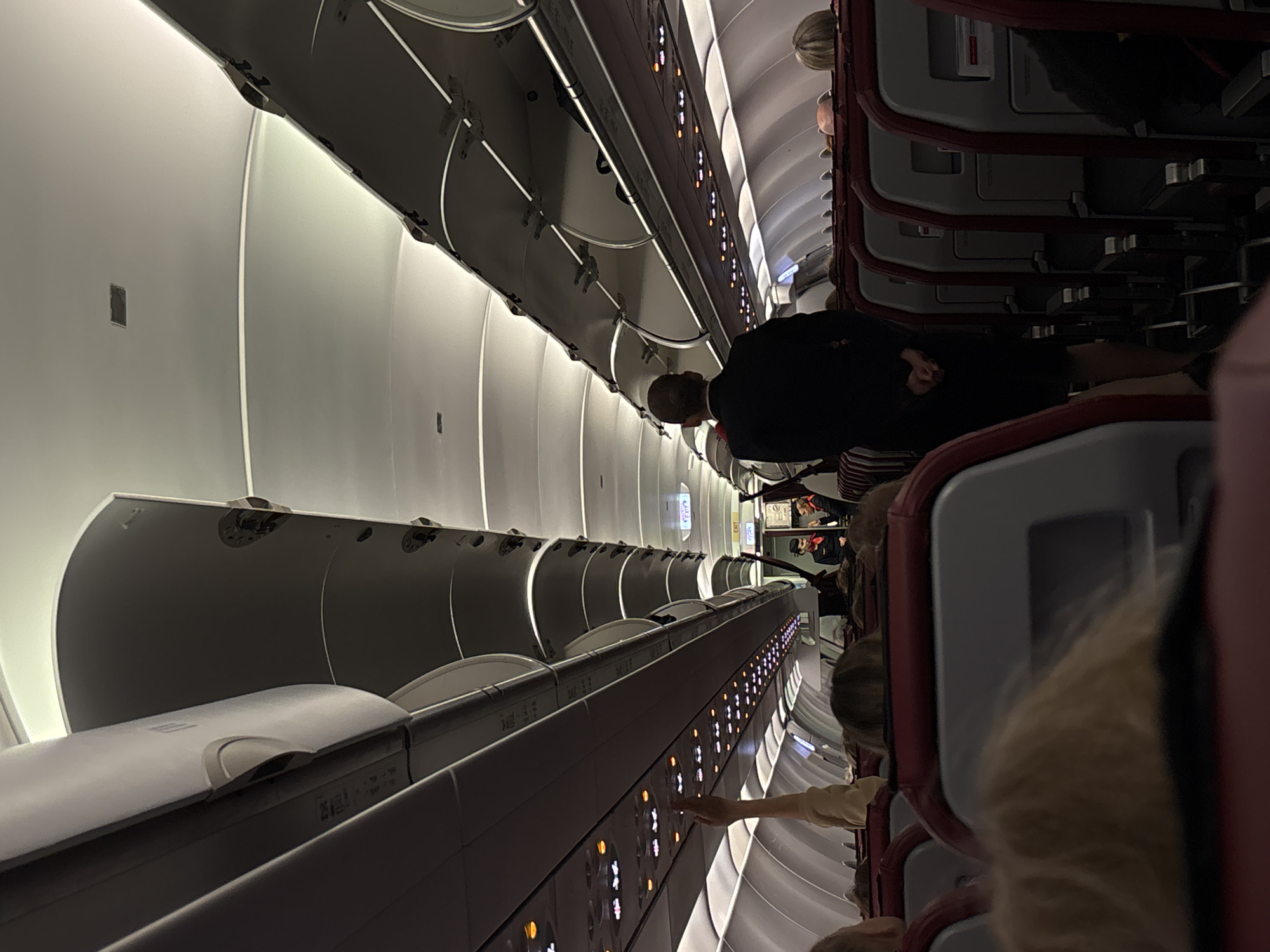 The image depicts the interior of an airplane cabin, likely on a commercial flight. The perspective is from a passenger seat looking forward towards the front of the aircraft. Overhead compartments are visible along both sides, as are rows of empty seats with red upholstery. Several passengers are seated, some with their heads visible. A flight attendant in a dark suit and red scarf stands in the aisle, facing away from the camera, with their hands clasped behind their back. Another flight attendant, wearing a similar uniform, is visible further down the aisle. The lighting is dim, with some illumination coming from overhead lights and indicator panels. There is no visible indication of the city of Bolshoye Savino, Russia, within the image itself. The scene is static, suggesting the plane is either preparing for or has just completed a flight.