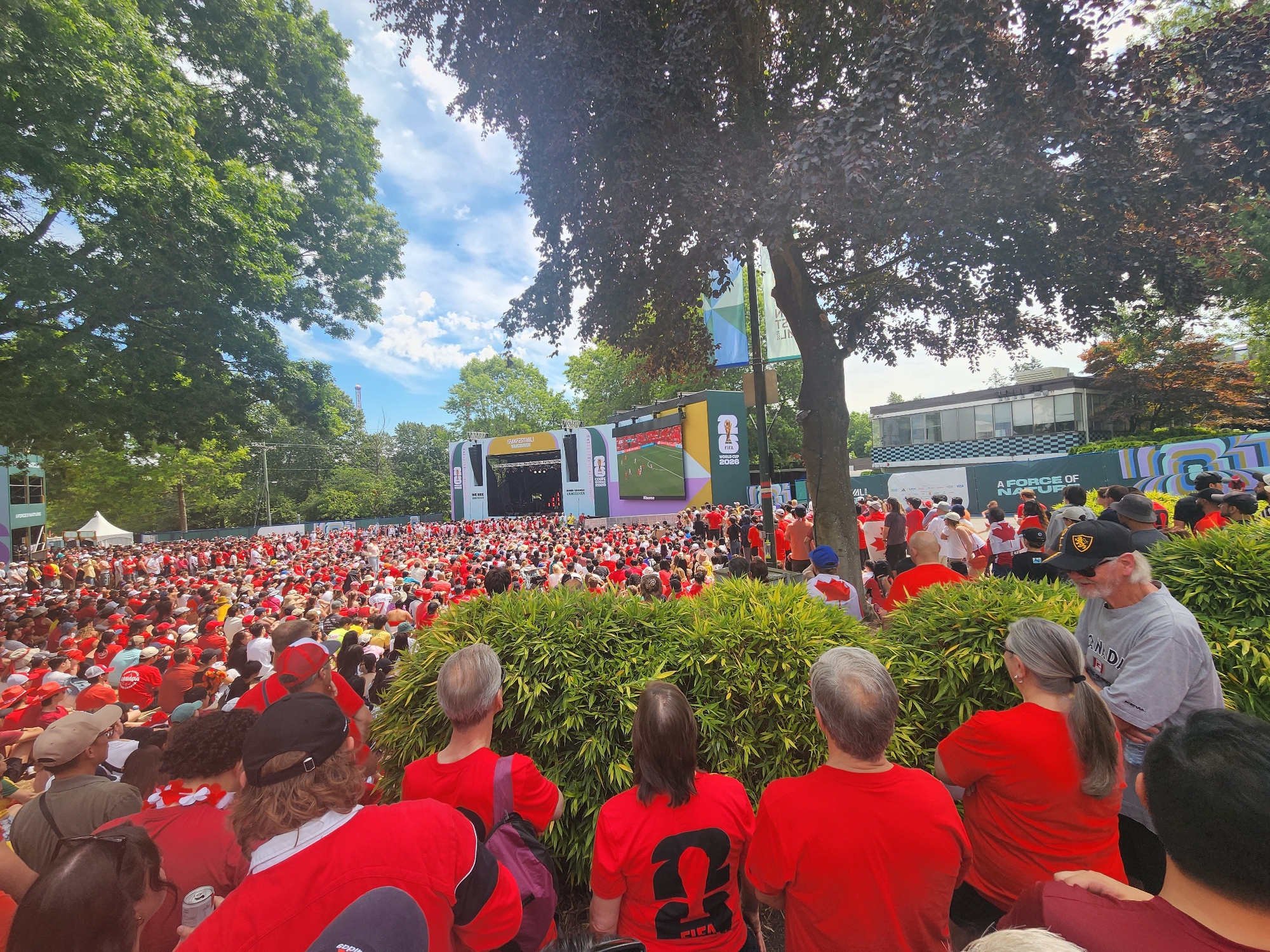 This media file captures a vibrant outdoor gathering of a large crowd, predominantly dressed in red, suggesting a shared allegiance or team support. The scene is dominated by a large screen displaying a soccer match, indicating the event is likely a public viewing of a sporting event, possibly the FIFA World Cup given the "FIFA WORLD CUP 2026" text visible on banners and the screen.
The setting appears to be a park or public square, with lush green trees framing the scene and providing shade. The weather seems pleasant, with a bright sky and scattered clouds, suggesting it's daytime. The crowd is dense, with people of all ages standing and sitting, some appearing to be very engaged with the match on the screen. The general atmosphere seems to be one of excitement and communal enjoyment.
Notable details include the large screen broadcasting the soccer game, with players visible on the field. Several banners with text are present, including "FIFA WORLD CUP 2026" and "A FORCE OF NATURE". The prevalence of red clothing and the Canadian flag designs on some individuals' attire suggest this might be a viewing party for a Canadian soccer team or a general celebration related to the World Cup in Canada. The emotions visible are a mix of focus on the game and cheerful engagement with fellow spectators. The overall impression is of a lively, communal event centered around a sporting spectacle.