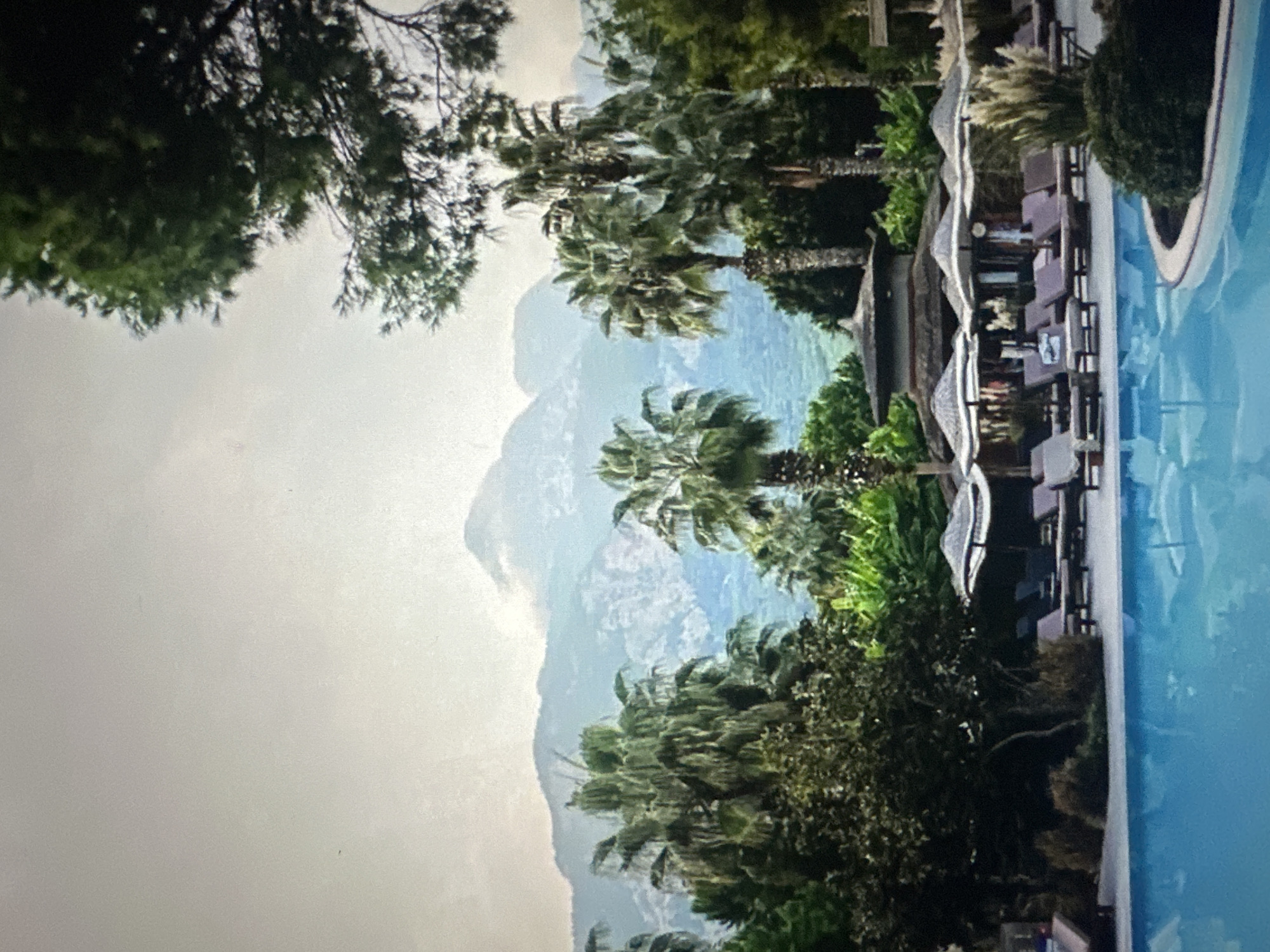 The image depicts a tranquil scene at a resort with a swimming pool in the foreground. The pool's surface reflects the sky and surrounding palm trees. Behind the pool, a paved area is furnished with lounge chairs and umbrellas, suggesting a relaxation area. A building with a thatched roof and several levels is visible behind the lounge chairs. A variety of palm trees and other lush vegetation dot the landscape, leading to a backdrop of majestic mountains shrouded in mist under an overcast sky. The overall impression is one of a serene, tropical getaway.