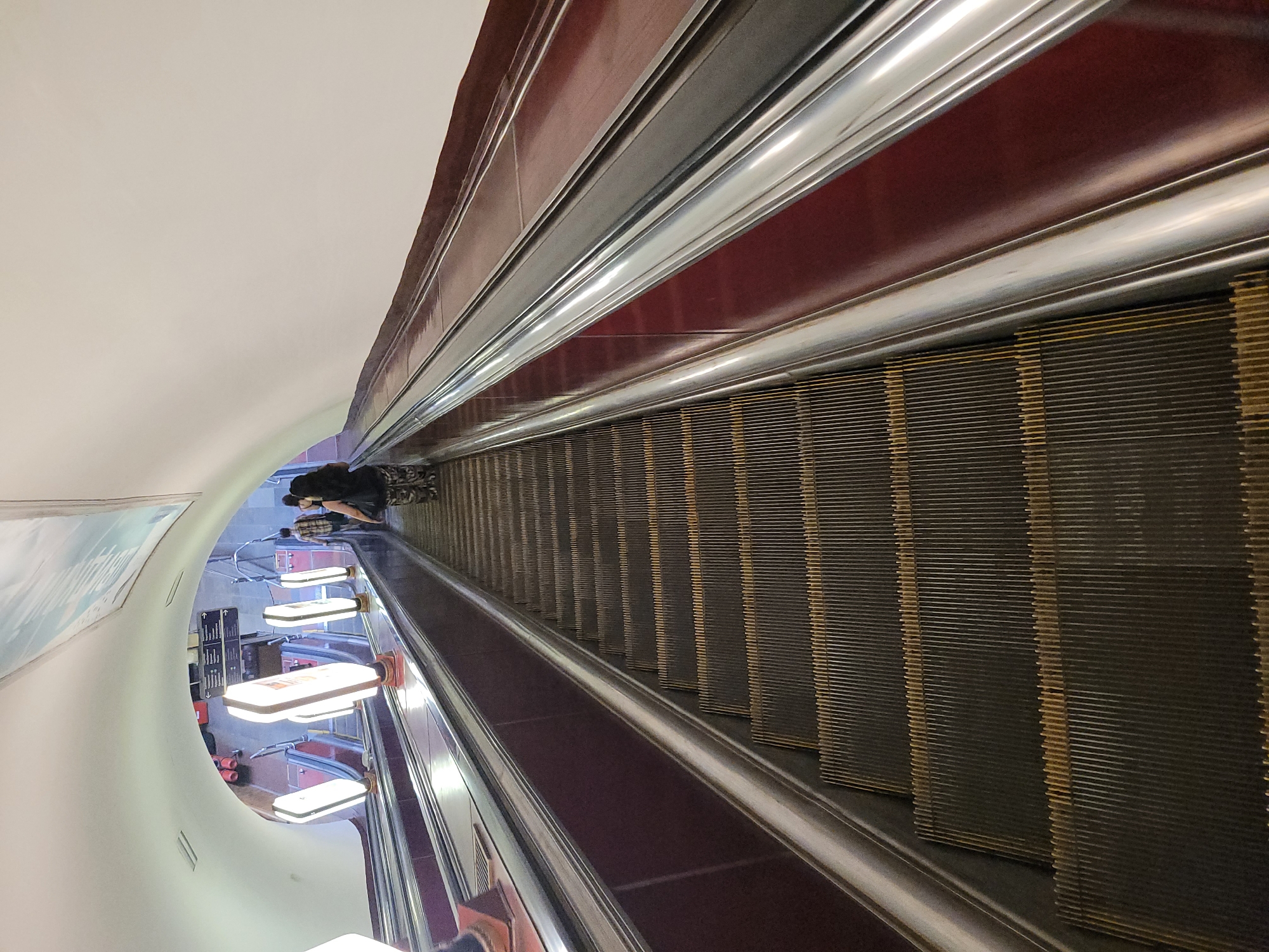 An upward-angled, full shot shows a wide escalator going up into a tunnel-like ceiling in the city of Unknown. The escalator steps are dark gray with yellow accents and are lined by a metallic handrail and a dark red side panel. The escalator is illuminated by large, glowing white rectangular lights on stands positioned along the side. In the distance, at the top of the escalator, two individuals are ascending. The person closest to the camera, a woman with dark hair, is wearing a dark top and patterned pants. Behind her, a man in a plaid shirt is visible. The tunnel ceiling is painted white and curves overhead, with some signage and lighting fixtures visible in the distance. The overall scene suggests a public transit station or a large building.