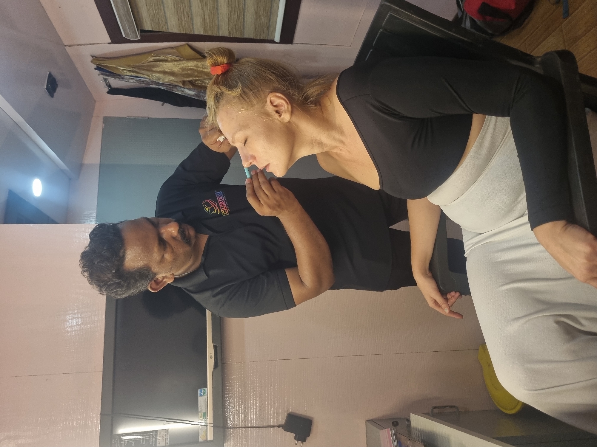 The image captures an indoor scene, likely in a salon or studio in Kalamassery, India, where a man is performing an activity on a seated woman.
The main subjects are a man and a woman. The man has dark, curly hair and a mustache, wearing a black long-sleeved shirt with a colorful logo. He appears to be focused on the task. The woman has light-colored hair pulled back with a red hair tie. She is wearing a black long-sleeved crop top and a light-colored, possibly white or light grey, form-fitting skirt or dress. She is seated on a dark chair, with her eyes closed and head slightly tilted, suggesting she is receiving a treatment or application.
The activity involves the man holding a small, light blue object (possibly an applicator or product dispenser) near the woman's face, specifically around her nose and upper lip. This suggests a beauty treatment, makeup application, or a similar cosmetic service.
The setting is an indoor room with light-colored, textured walls. In the background, a black television screen with "LG" branding is visible, along with cables and a power adapter. Garments or fabrics are hanging on a rack or hook in the upper background. A dark table or counter is positioned next to the woman.
Notable details include artificial lighting, typical of an indoor space. The woman wears a ring on her right hand. The man's shirt features a prominent logo that reads "CUBES ENTERTAINMENT" with a graphic of interconnected cubes. The television also displays a green energy efficiency sticker. The atmosphere appears calm and professional, with both individuals focused on the ongoing process.