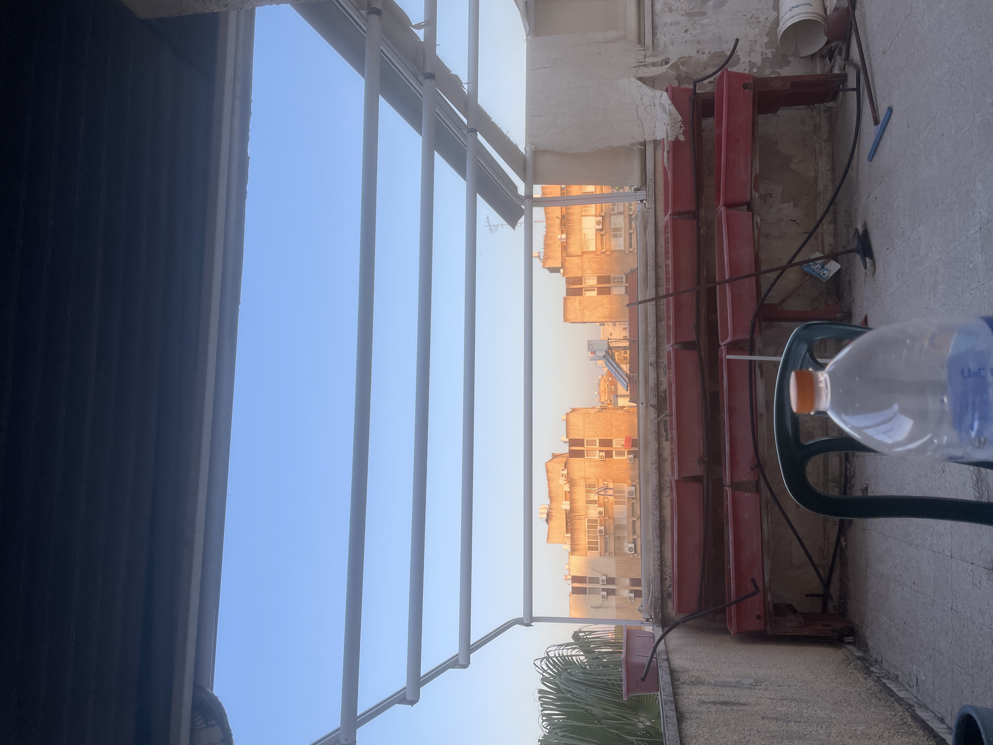 The image depicts an urban landscape viewed from a balcony or terrace, oriented as if looking out from under an overhang. In the immediate foreground on the left, dark, louvered shutters or an awning create a shaded area. On the right, a concrete or tiled floor is visible, featuring a green plastic chair and a clear plastic water bottle with an orange cap, where the text "U-C" is partially legible.
Further back on the right, a red metal shelving unit, possibly a planter stand, rests against a textured, light-colored wall. Above this unit, an upside-down white plastic bucket with the word "tambur" (likely "Tambour") printed on it is visible, along with a blue broom head and some cables on the floor.
A series of white vertical bars, suggesting a balcony railing or pergola, frame the mid-ground. Beyond these bars, the horizon reveals a cityscape under a clear, bright sky. Distant multi-story buildings, mostly light brown or beige, are illuminated by the warm, golden light typical of either early morning or late afternoon (golden hour). Numerous external air conditioning units are visible on these buildings. Green palm fronds are also visible near the bottom right of the distant view. The scene is static, showing no direct activity, and conveys a calm atmosphere in an urban setting within Petah Tikva, Israel.