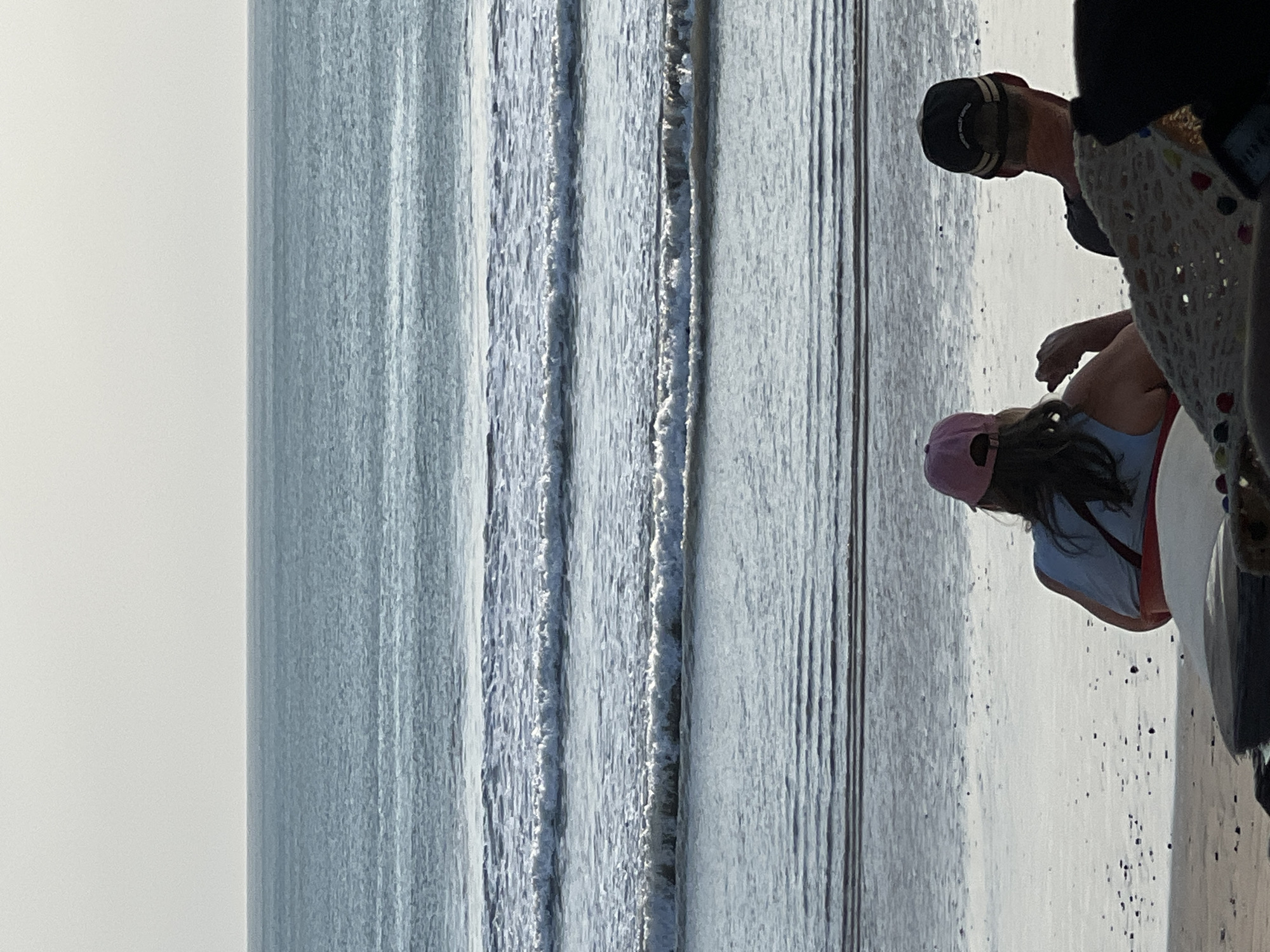 The image shows two people from behind, sitting on a beach and looking out at the ocean. The person on the left is wearing a pink baseball cap and a light blue top, with long dark hair. The person on the right is wearing a dark baseball cap and a dark t-shirt. They are both seated on the sand. The ocean is calm with gentle waves rolling towards the shore, creating horizontal lines across the water. The sky is overcast and a pale grey. The beach is sandy and appears to be wet closer to the water. The scene suggests a relaxed atmosphere, possibly a sunset or early morning view. This scene is typical of the Kuta beach in Bali, Indonesia, known for its long stretches of sand and surf.