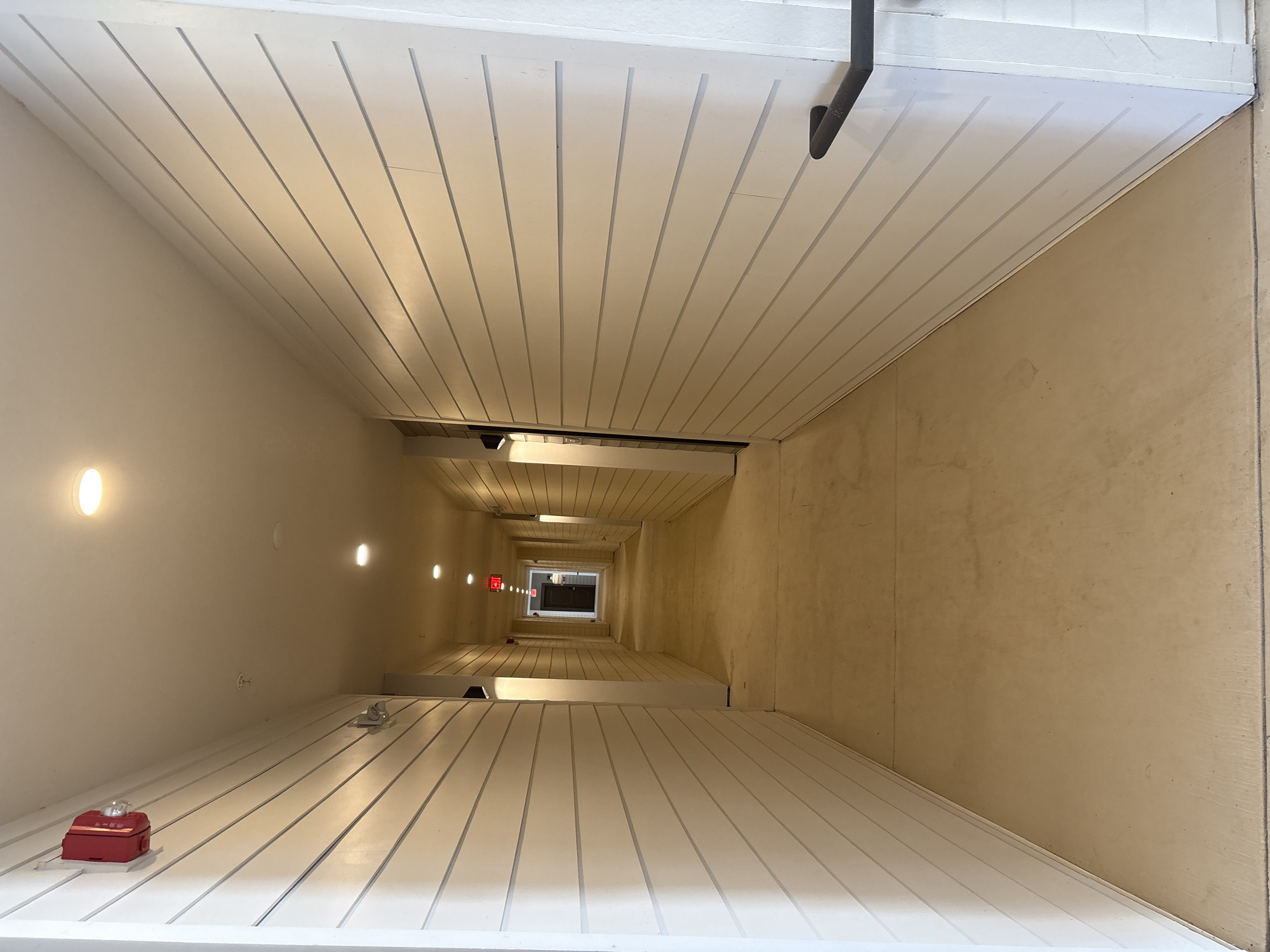 The image shows a long, brightly lit hallway with white shiplap walls and a light beige concrete floor. Recessed ceiling lights illuminate the space. On the left wall, a red fire alarm is mounted high up. Further down the hallway, an "EXIT" sign with red lettering is visible. The hallway appears to be in a building in Palmetto, United States. There are no people or animals visible in the image. The hallway is clean and well-maintained, with no visible damage or disarray. The perspective of the image is from the ground level, looking down the length of the hallway towards a door at the far end.