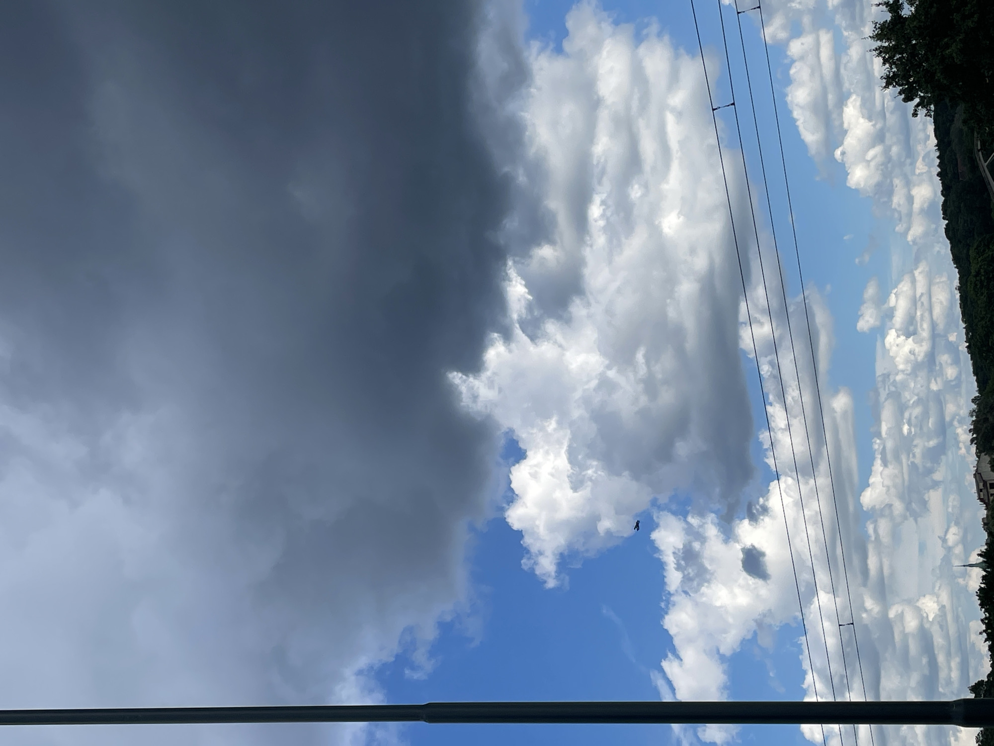 A low-angle outdoor shot shows a dark cloud formation overhead, partially obscuring a bright blue sky dotted with white cumulus clouds. A tall, dark pole stands on the left side of the frame, running vertically from top to bottom. Several black wires, presumably from an overhead power line, stretch horizontally across the middle of the image. In the lower half of the frame, a lush green landscape with trees and buildings is visible under the scattered clouds. A small, dark object, possibly a drone or a distant airplane, is visible in the upper-middle section of the frame against the blue sky. The overall lighting suggests a partly cloudy day.