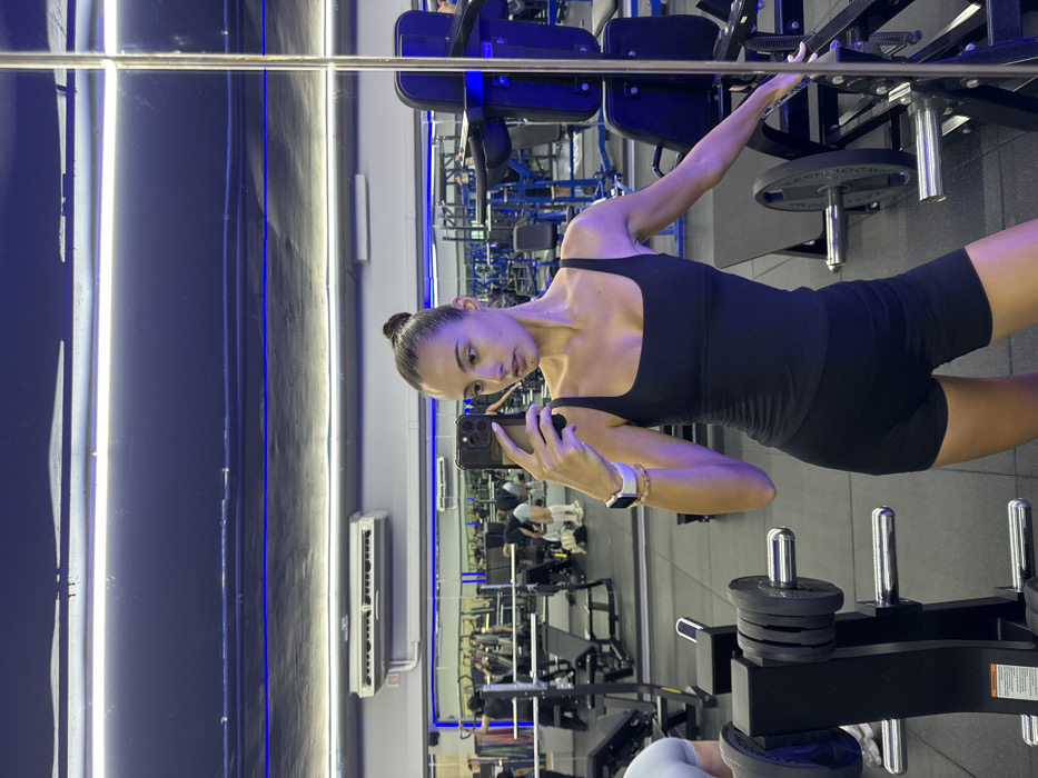 A woman is depicted taking a mirror selfie inside a gym located in Bangkok, Thailand. She stands in the foreground, looking slightly to her left, holding a dark-colored smartphone in her left hand to capture her reflection. She is wearing a black square-neck tank top and black athletic shorts, with an Apple Watch visible on her left wrist. Her hair is pulled back into a bun. Her right arm is extended, resting on a piece of dark-colored gym equipment. The gym environment features dark walls, a dark ceiling, and a dark rubberized floor. The space is illuminated by overhead white strip lights and blue accent LED strip lighting along the walls. In the background, reflected in the large wall mirror, various gym equipment including weight racks, benches, and exercise machines are visible. Several indistinct individuals are also seen in the reflection, engaged in exercise. A white wall-mounted air conditioning unit is reflected on the upper left wall. A black weight plate storage rack with chrome posts is partially visible in the left foreground.