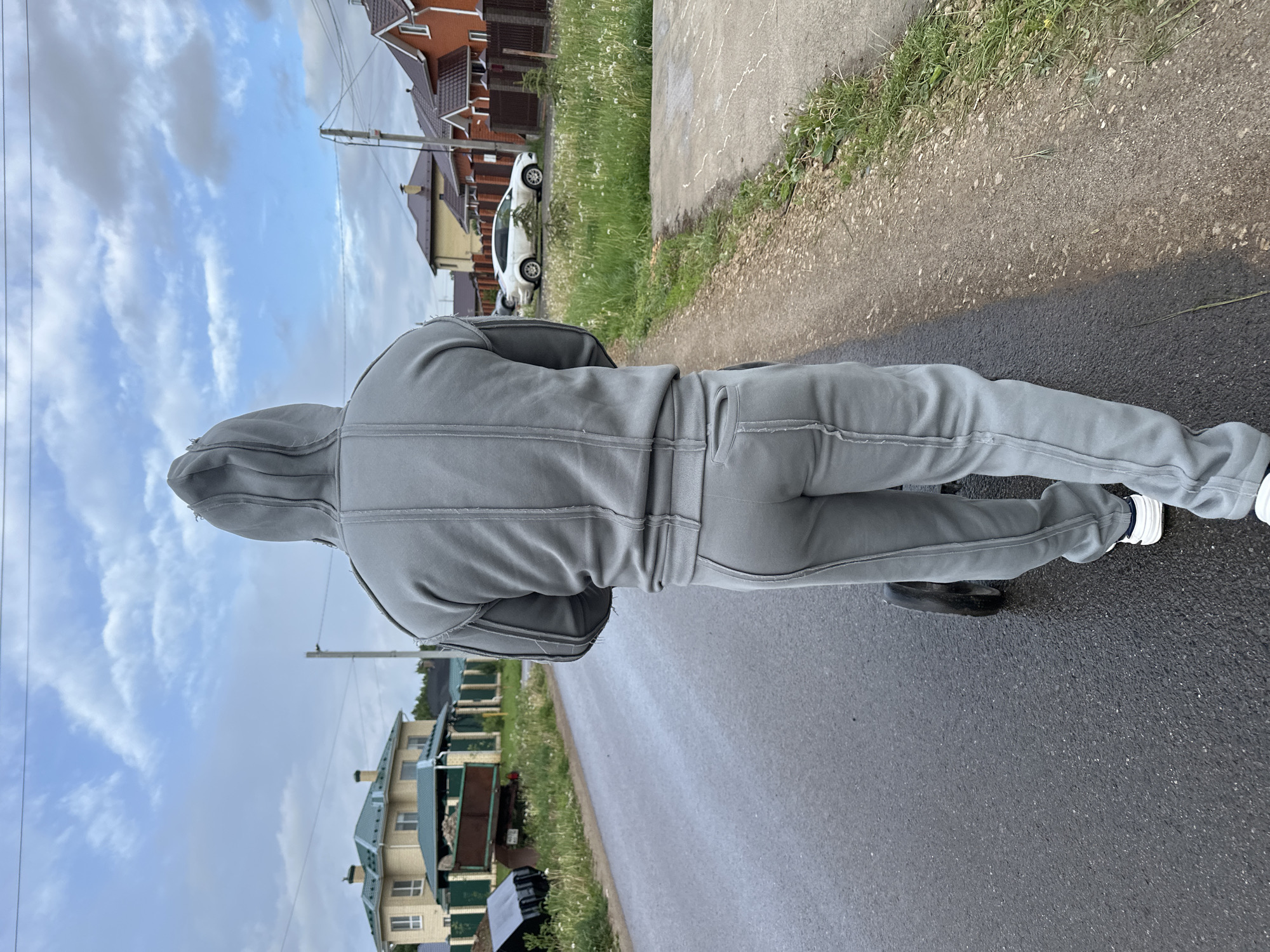 A person wearing a gray hooded tracksuit is walking along a paved road in a residential area of Эстачи, Russia. The road appears wet, and houses with varying architectural styles are visible in the background, with some having brown roofs and others with green roofs. A white car is parked near one of the houses. Vegetation lines the side of the road and separates the pavement from the residential properties. The sky is partly cloudy.