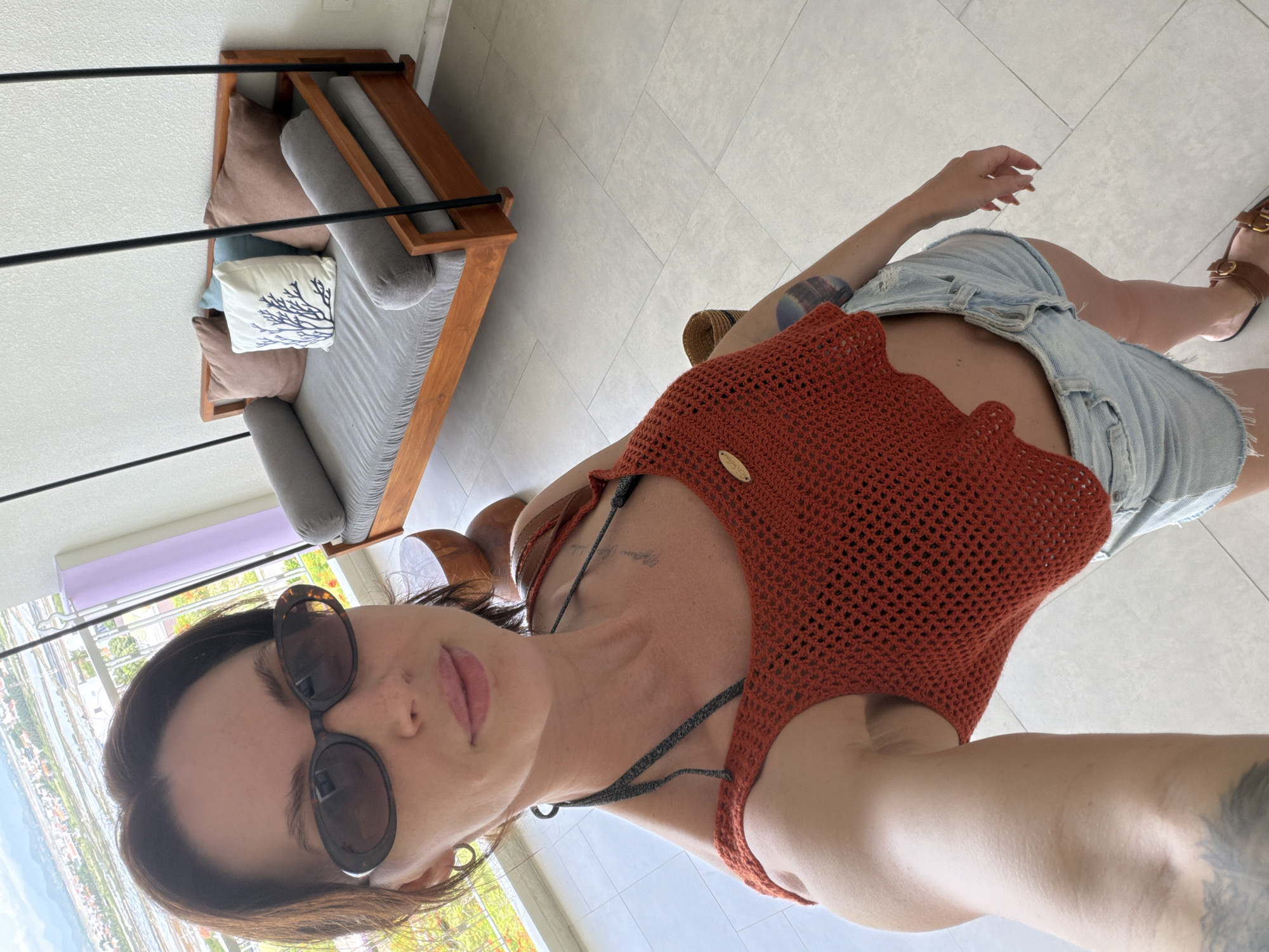 In a bright, indoor setting, a woman with brown hair and sunglasses takes a selfie. She is wearing an orange crocheted crop top and light blue denim shorts. Behind her, a wooden swing daybed with grey cushions and decorative pillows is visible against a white wall. The floor is tiled in a light, neutral color. Through a balcony railing in the background, a distant view of a city and body of water can be seen, suggesting a location in a coastal area. The scene appears to be within a residential or hospitality space. The image is taken from a high angle, looking down at the woman.