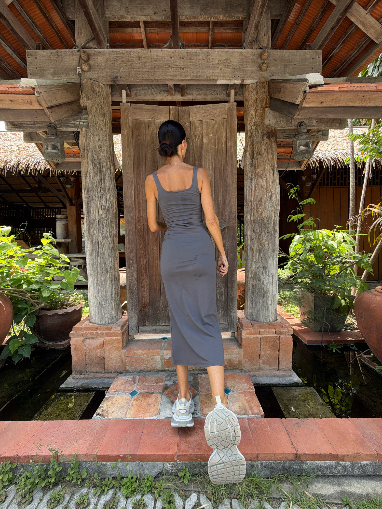 A woman stands with her back to the camera, wearing a grey tank dress and white sneakers, on a brick pathway in front of a weathered wooden structure. The structure appears to be a building with a thatched roof and wooden beams, reminiscent of traditional Southeast Asian architecture, possibly located in Koh Samui District, Thailand. Lush green plants in terracotta pots surround the pathway, and a small, dark body of water is visible in the foreground. The overall setting suggests an outdoor, possibly tropical, environment.