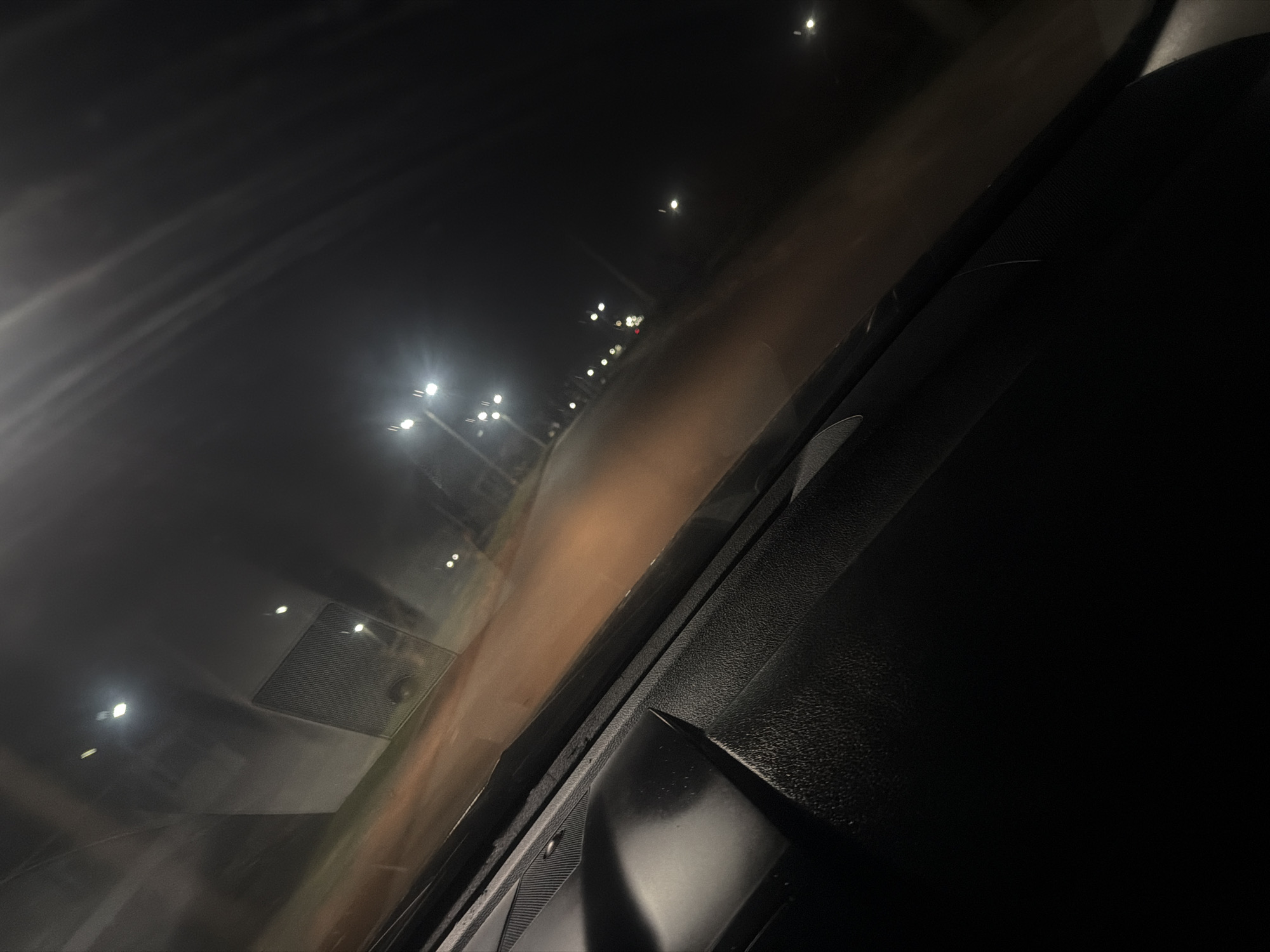 The image depicts a view from inside a vehicle at night, likely a car. The interior dashboard is visible, made of textured black plastic. Streetlights line the road outside, reflected in the windshield. The perspective suggests motion, as if the vehicle is traveling along a road illuminated by these lights. There is no evidence to conclusively place the image in Iddo Tudunwada, Nigeria. No people are visible.