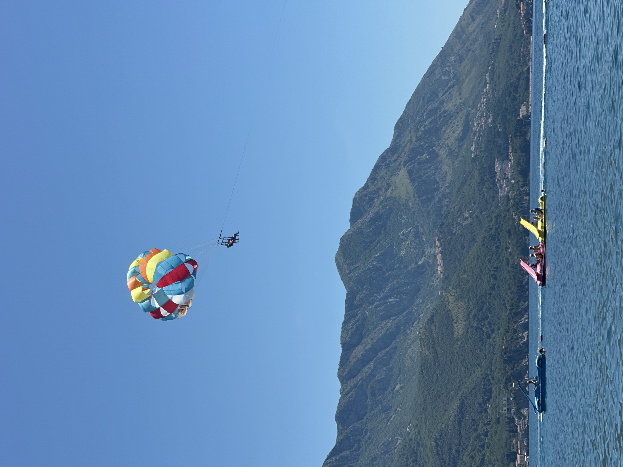 In Boreti, Montenegro, on a clear, sunny day, a colorful parasail drifts across a bright blue sky, carrying two individuals suspended beneath it. Below, the deep blue Adriatic Sea is dotted with three colorful watercraft. To the left, a blue boat with a single occupant cuts through the water. To its right, a pink watercraft, possibly a paddleboat with a slide, carries several passengers. Further to the right, a yellow watercraft, also appearing to be a paddleboat, has two people on board. The background features a majestic, tree-covered mountain range, with scattered buildings visible along its slopes and at its base. The scene depicts a vibrant summer day filled with recreational activities on the water.
