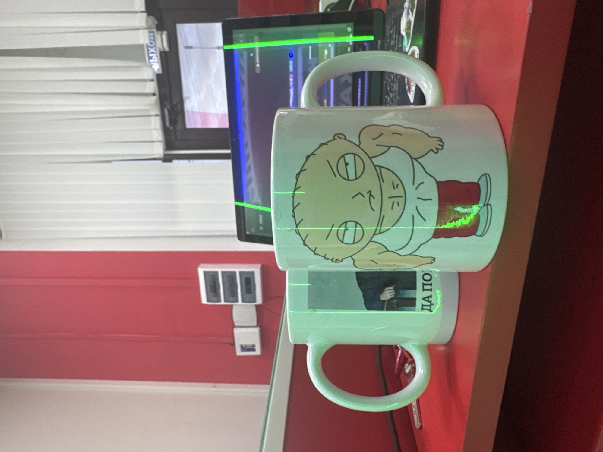 The image depicts two mugs on a red table. The mug in the foreground features a cartoon character with an angry expression, muscular arms, and is wearing a white tank top and red pants. The mug to the left is lighter in color and has a photo or graphic of a person's arm and hand resting on a dark surface. Green laser lines are visible crossing both mugs and a laptop screen in the background. The background shows a doorway with vertical blinds and a sign that reads "ВЫХОД" (EXIT in Russian). Electrical boxes are mounted on a red wall to the left. The scene is indoors, possibly an office or a room with a desk. The location of Yegorlykskaya, Russia is not evident from the visual information in the image.