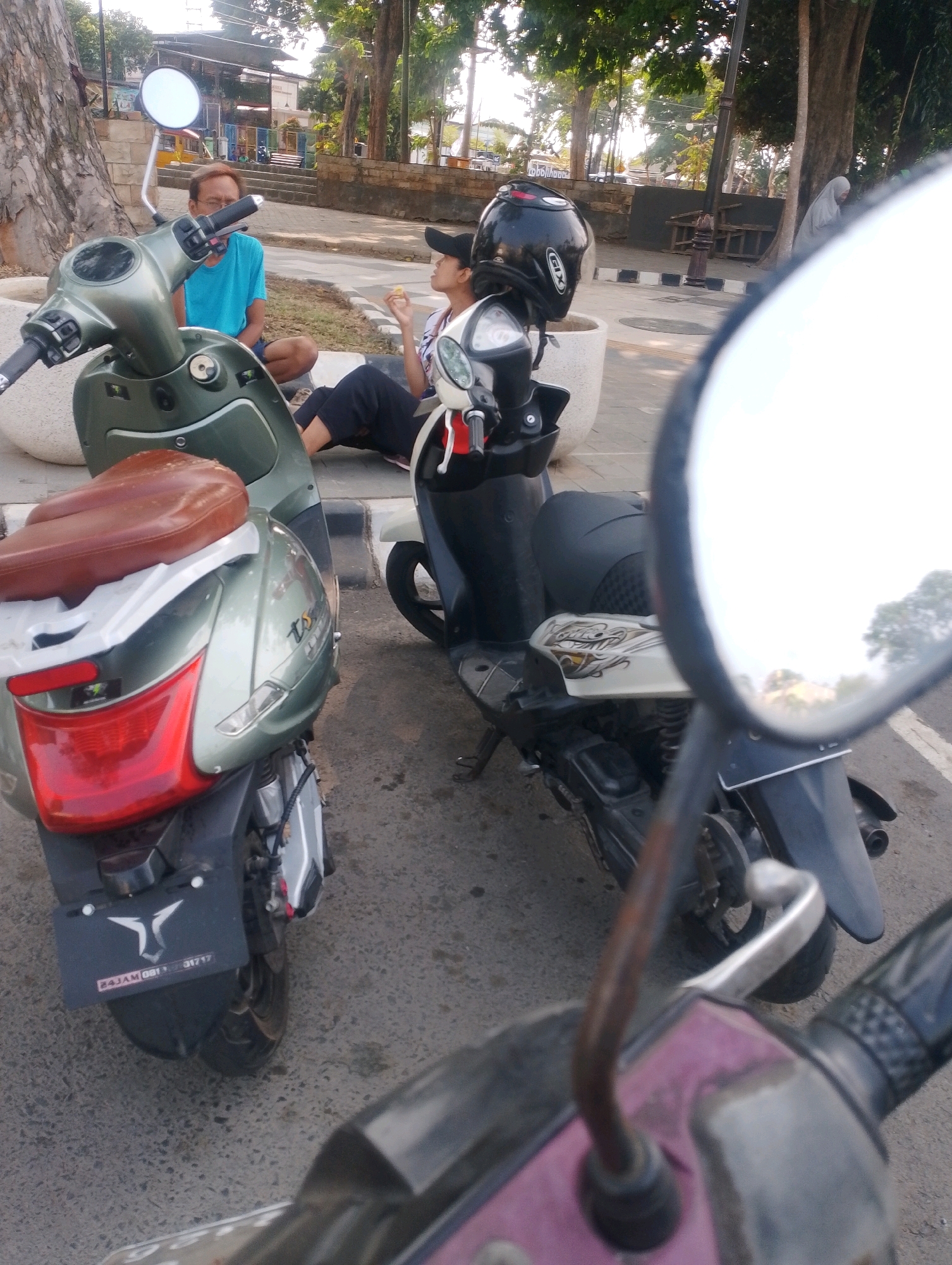 Two scooters are parked on a paved surface. In the foreground, a green scooter is partially visible from the rear, showing its red taillight and a license plate with "24JAM 001 01717" and a logo. Behind it, a black scooter with a black helmet on its handlebars is parked. To the left, a man wearing a blue t-shirt and shorts is seated on the ground, looking towards the right. To his right, a person in black pants and a white and pink shirt is also seated on the ground, holding a yellow object to their mouth. The background shows trees, a building, and what appears to be a sidewalk with planters. The scene suggests a public outdoor space in Probolinggo, Indonesia, possibly a park or street side.