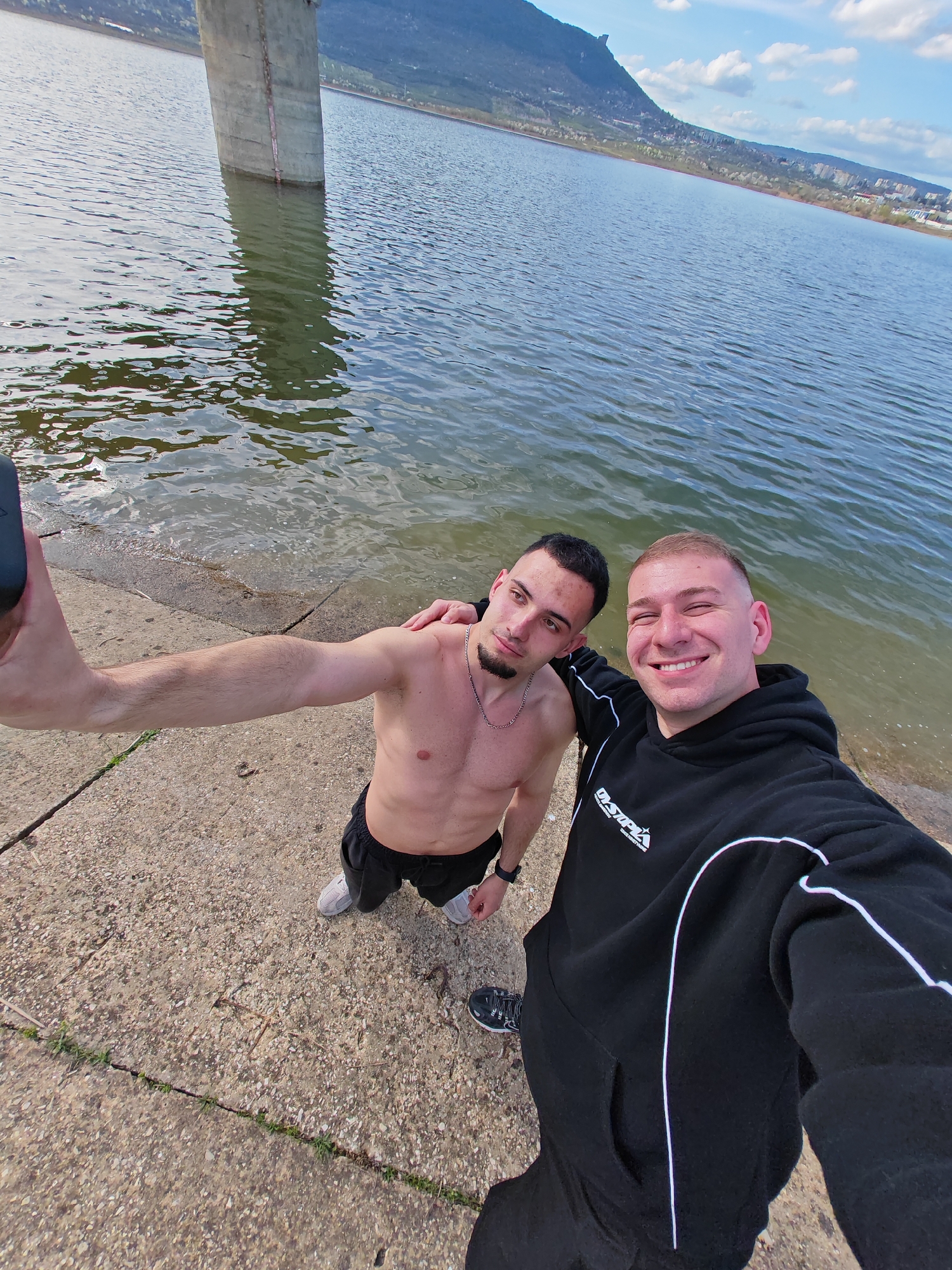 Two men stand on a concrete bank next to a body of water. The man on the left is shirtless, wearing shorts and sneakers. He has dark hair and a goatee, and is extending his arm to hold a black phone for a selfie. The man on the right is wearing a black hoodie and sweatpants with a white stripe down the side. He has short blonde hair and is smiling at the camera. In the background, there is a concrete structure, likely a support for a bridge or dam, and a mountain range under a blue sky with scattered clouds. The location is near the city of Shumen, Bulgaria, given the landscape and the style of the concrete structure. The men are likely taking a selfie near a lake or reservoir.