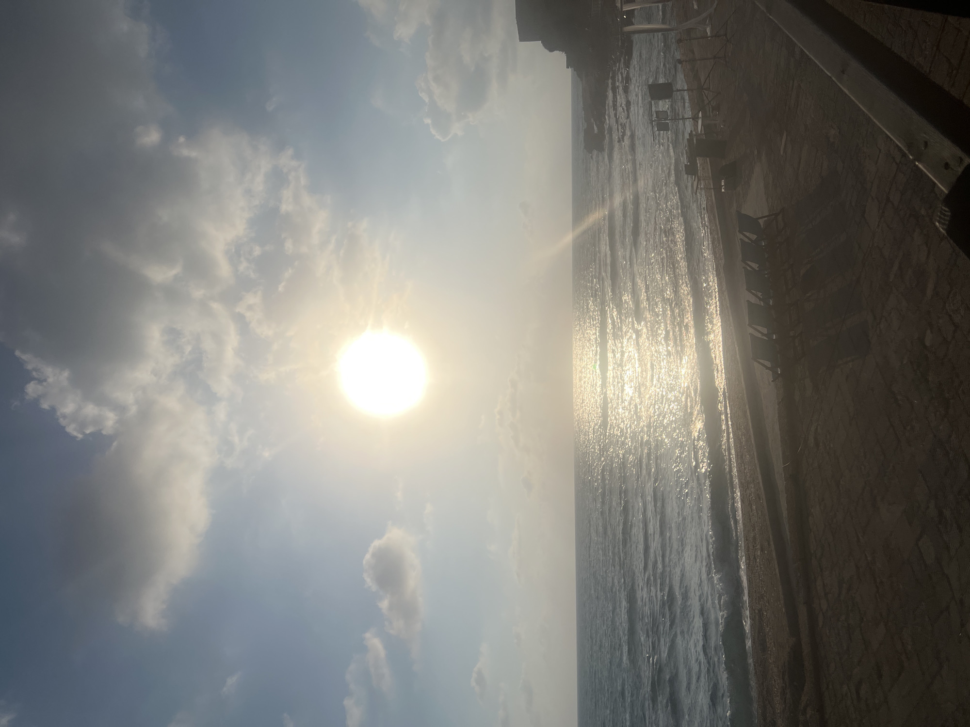 The image depicts a beach scene in Hof HaCarmel Regional Council, Israel, with a bright sun low in the sky, casting a reflection on the water. The sea is calm with gentle waves lapping at the shore. The beach is sandy, with a paved area in the foreground that appears to be set up for an event, featuring rows of chairs, a table with equipment, and speakers on stands. In the distance, a rocky coastline is visible with a structure on top. The sky is partly cloudy, with the sun partially obscured by clouds.
