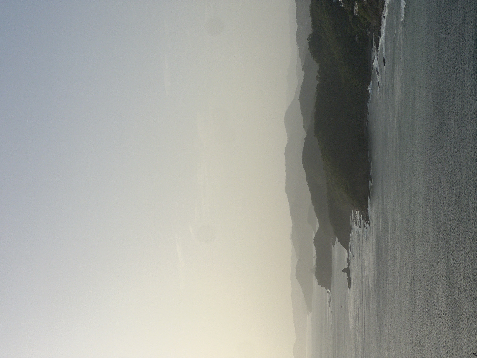 This is a landscape photograph of a hazy coastline with layered mountains in the distance. The foreground is dominated by the ocean, with textured, dark grey water that shows subtle wave patterns. Along the middle ground, a rugged, green-covered coastline juts out into the sea. Small white waves crash against rocky outcrops and the shore. In the far distance, several layers of mountains are visible, fading into a pale, hazy sky that transitions from a light grey at the top to a yellowish hue towards the horizon. A few small, white structures are sparsely visible on the upper slopes of the closest mountains to the right, suggesting some development. No people or explicit actions are depicted in the scene. The overall impression is one of a serene, possibly early morning or late afternoon coastal view. While the image depicts a coastline, it is not possible to pinpoint the exact location within San Juan del Sur, Nicaragua, based solely on the visual information provided.