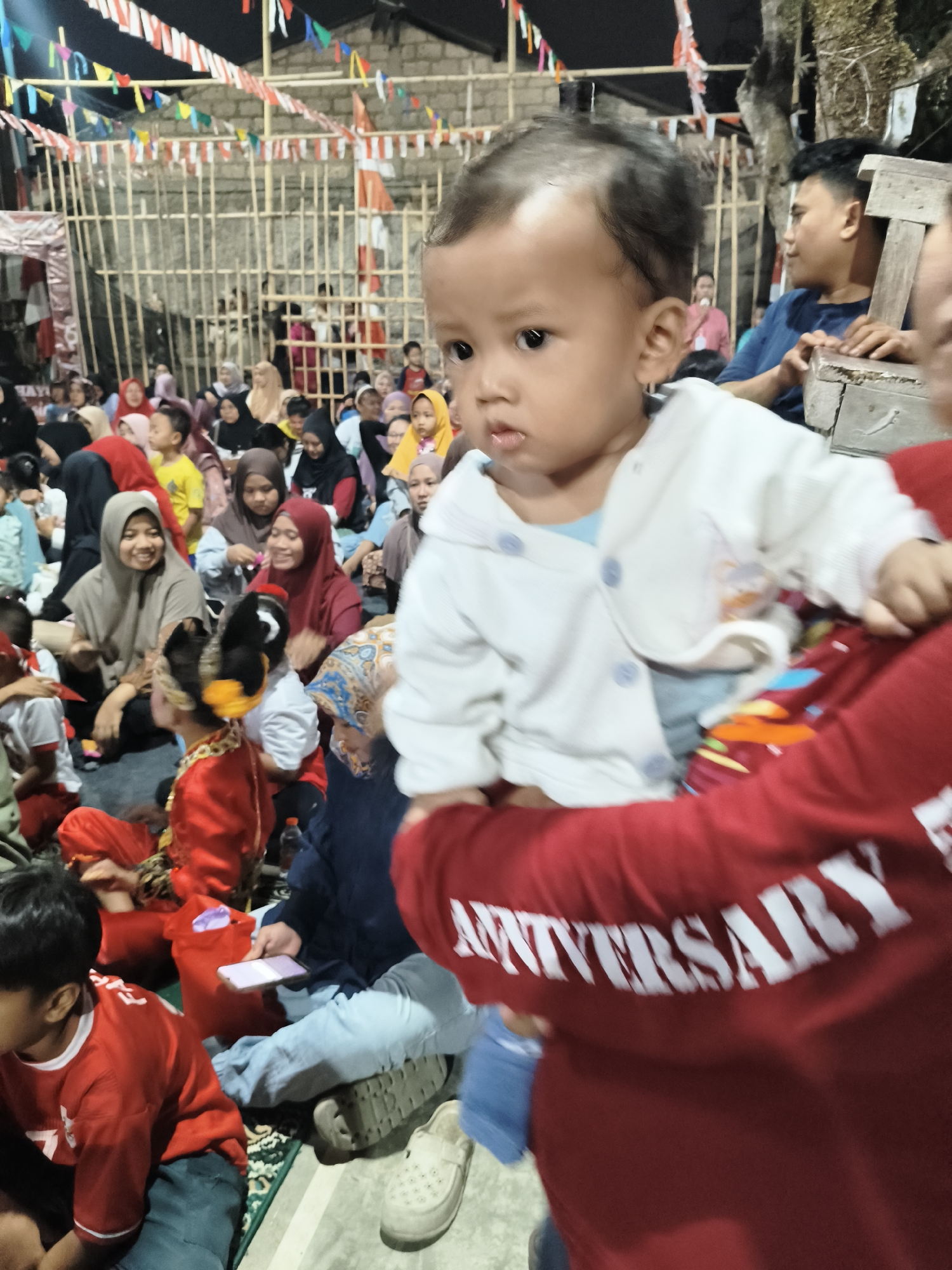 The image captures a close-up of a baby being held in the foreground, with a blurred background of a gathering of people. The baby is wearing a white jacket and appears to be looking slightly to the left. Behind the baby, a person in a red shirt with the word "ANNIVERSARY" visible is partially in view, holding the baby.
The background reveals a crowd of people, many of whom are wearing headscarves and traditional attire. The setting appears to be an outdoor event, possibly a celebration or community gathering, with bamboo fencing and strings of colorful flags overhead, suggesting a festive atmosphere. There are also some structures visible, including what looks like a brick wall. The lighting suggests it is evening or night.
While the image does not explicitly state the location, the mention of "South Tangerang, Indonesia" provides context for the cultural attire and general ambiance depicted. The scene suggests a local event within a community in South Tangerang, Indonesia.