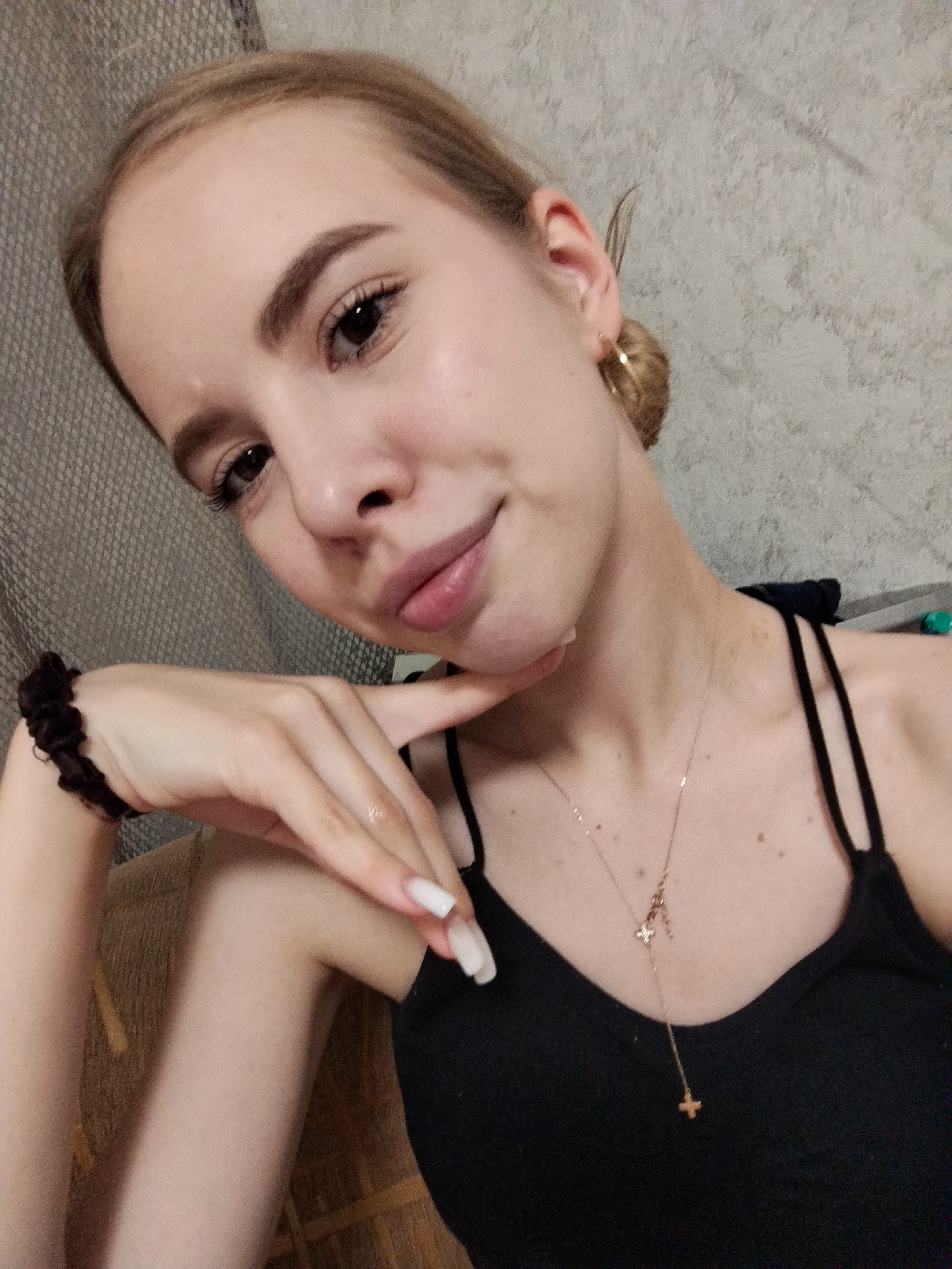 A young woman with blonde hair pulled into a bun is shown from the shoulders up, angled towards the viewer. She is wearing a black tank top with thin straps and a long necklace with two small cross pendants. Her left hand rests on her chin, with long white acrylic nails. A dark scrunchie is visible on her left wrist. The background consists of a textured gray wall and a brown couch.