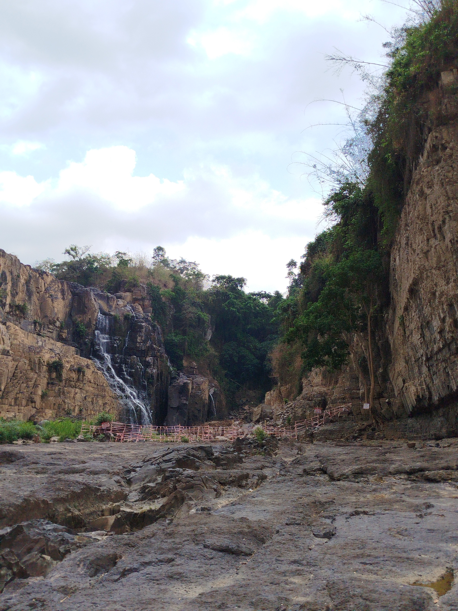 The image depicts a waterfall and rocky landscape, located in Tân Phú, Vietnam. The foreground is dominated by a large expanse of dark, cracked rock. A flimsy red and white barrier is strung across the midground, preventing access to the area directly below the waterfall. The waterfall itself cascades down a cliff face on the left side of the image. Lush greenery surrounds the waterfall and grows on the cliffs on either side of the scene. The sky is overcast with thick clouds.